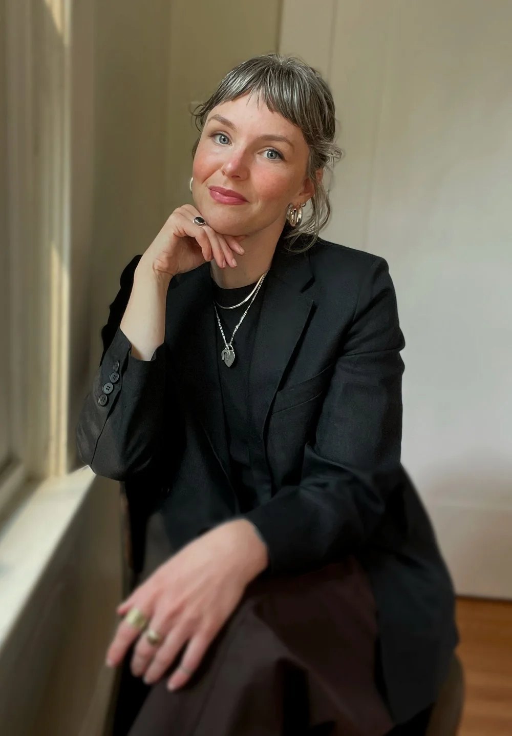 A woman with short gray hair, light skin, and blue eyes sitting near a window, wearing a black blazer and jewelry, resting her chin on her hand and smiling at the camera.