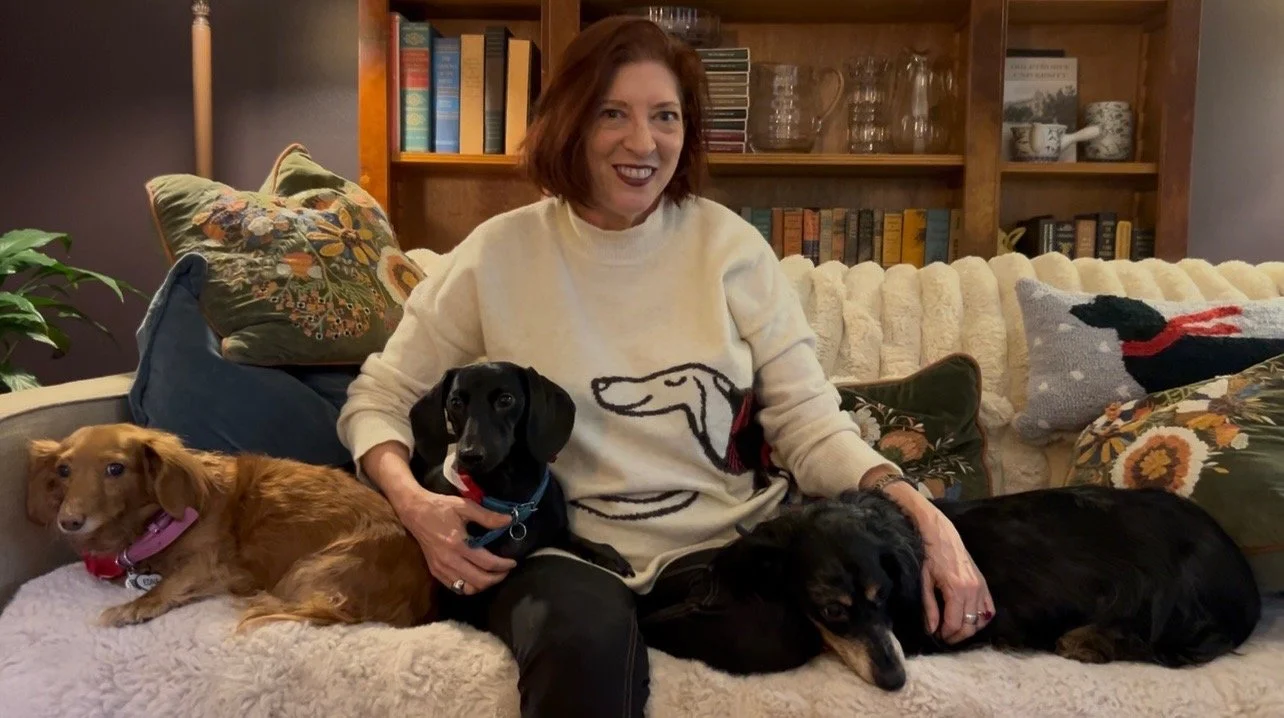 A woman with red hair smiling and sitting on a couch surrounded by three dogs in a cozy living room. The woman is wearing a cream sweater with a dachshund outline on it. The background has wooden shelves with books and glassware, and decorative pillows.