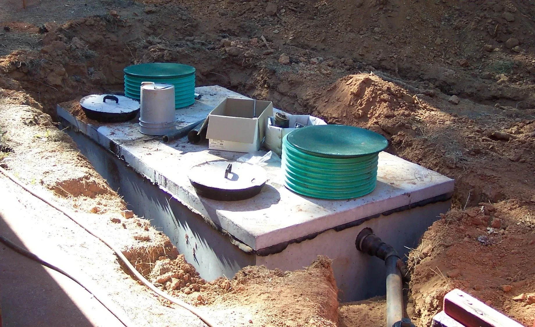 Construction site with a rectangular concrete foundation and utility pipes, with red dirt surrounding it, in an outdoor setting.