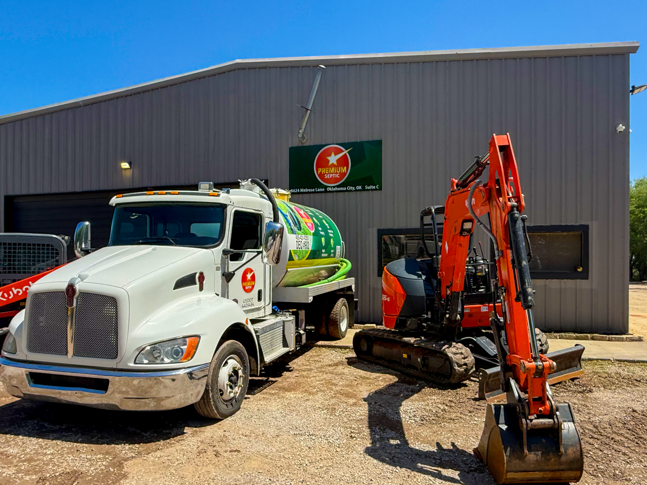 A white septic truck with a green septic tank attached, parked next to an orange mini excavator with tracks, outside a gray industrial building with a sign that says 'Premium Septic'.
