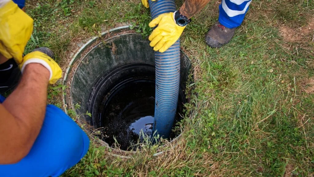 People working around an open underground well or septic tank, inserting or removing a blue corrugated pipe, wearing yellow gloves.