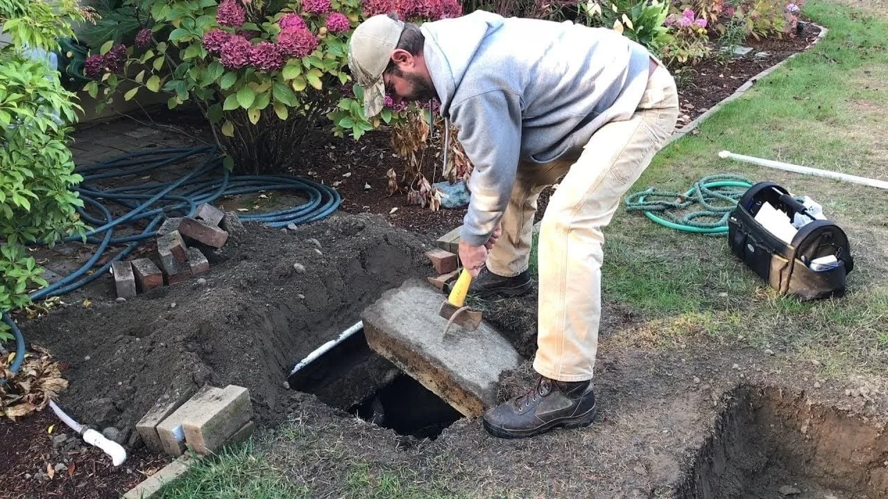 A man working in a garden, lifting a large concrete slab from a hole in the ground with a hammer and chisel, surrounded by gardening tools and hoses.