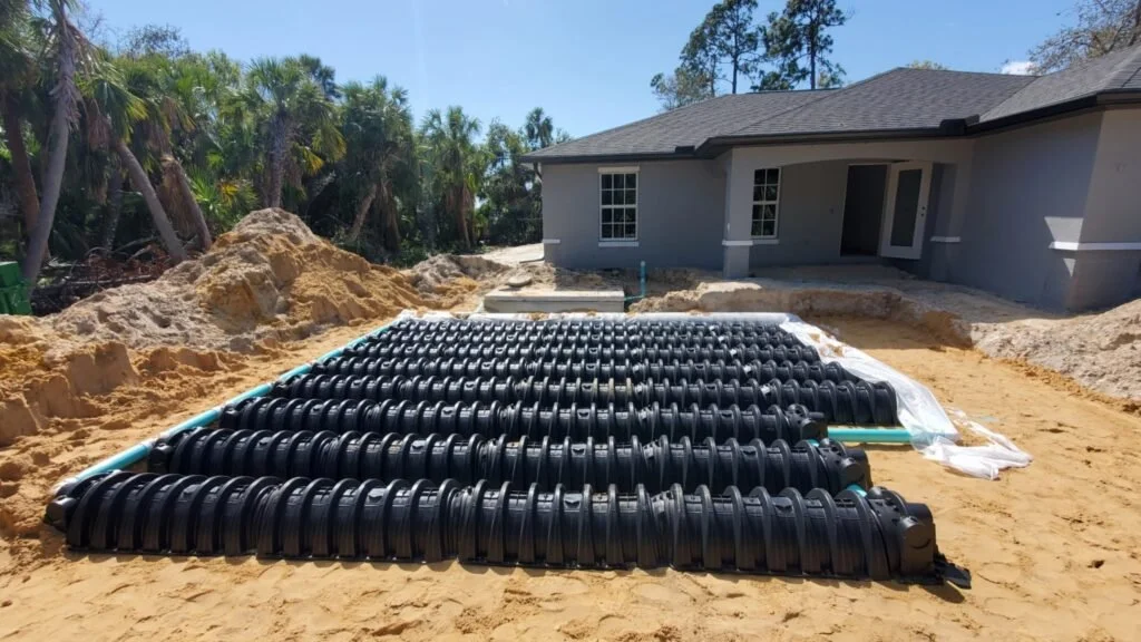 Installation of a drainage system with black plastic pipes and corrugated fittings in a backyard construction site near a gray house surrounded by palm trees.