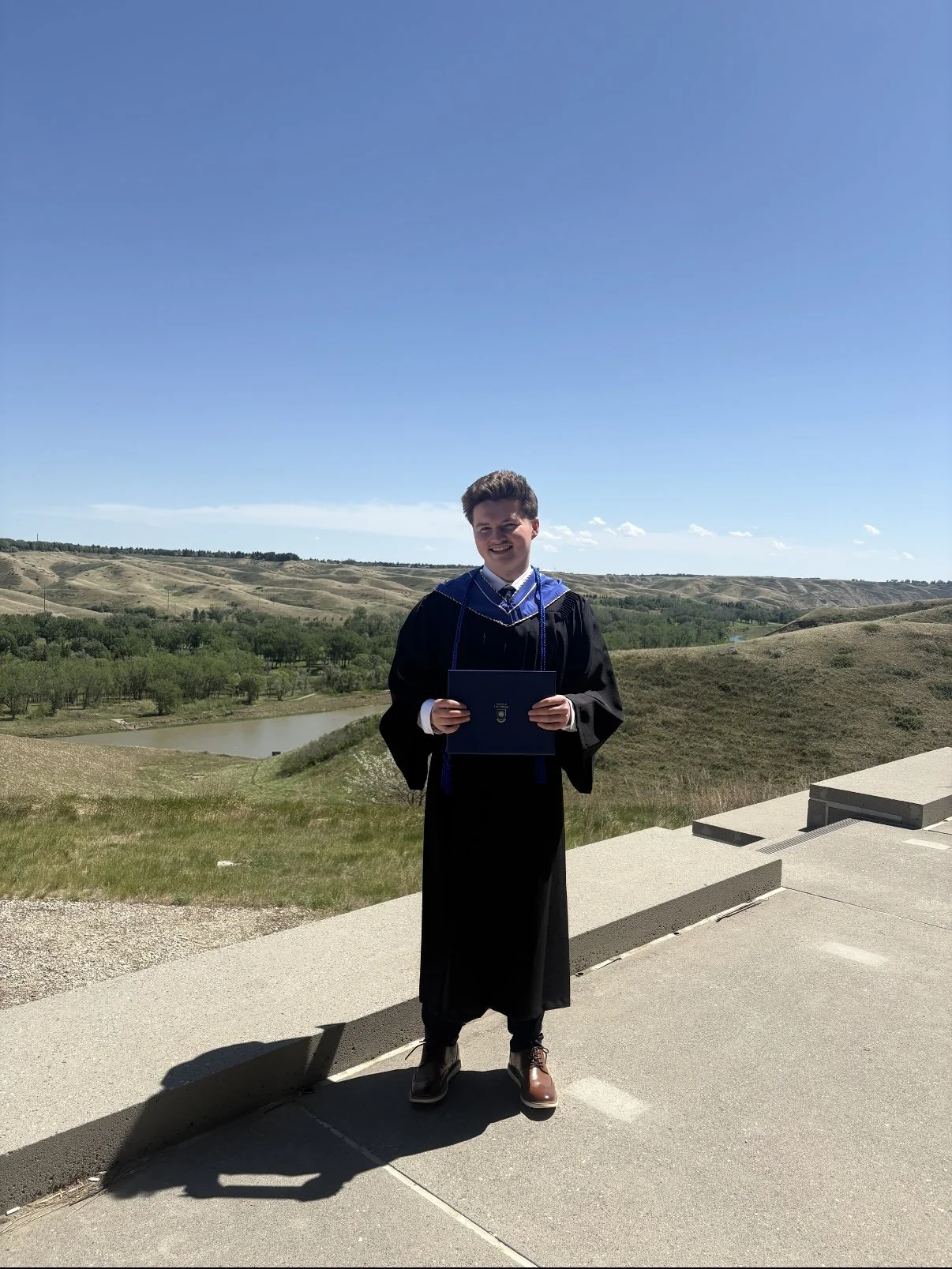 A young man in graduation attire holding a diploma, standing outdoors on a concrete platform with rolling hills and a river in the background under a clear blue sky.