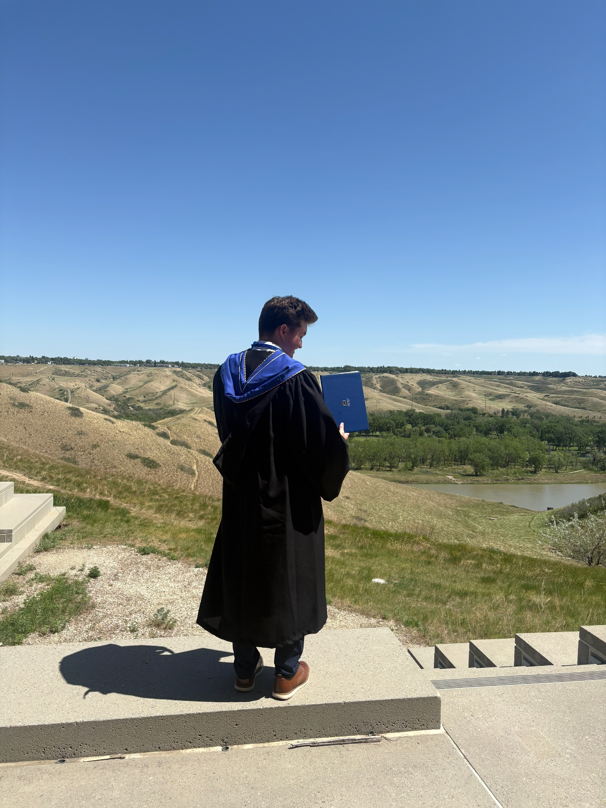 A young man in a graduation gown and blue stole stands on steps outdoors, holding a diploma folder, overlooking a hilly landscape with a river and trees under a blue sky.