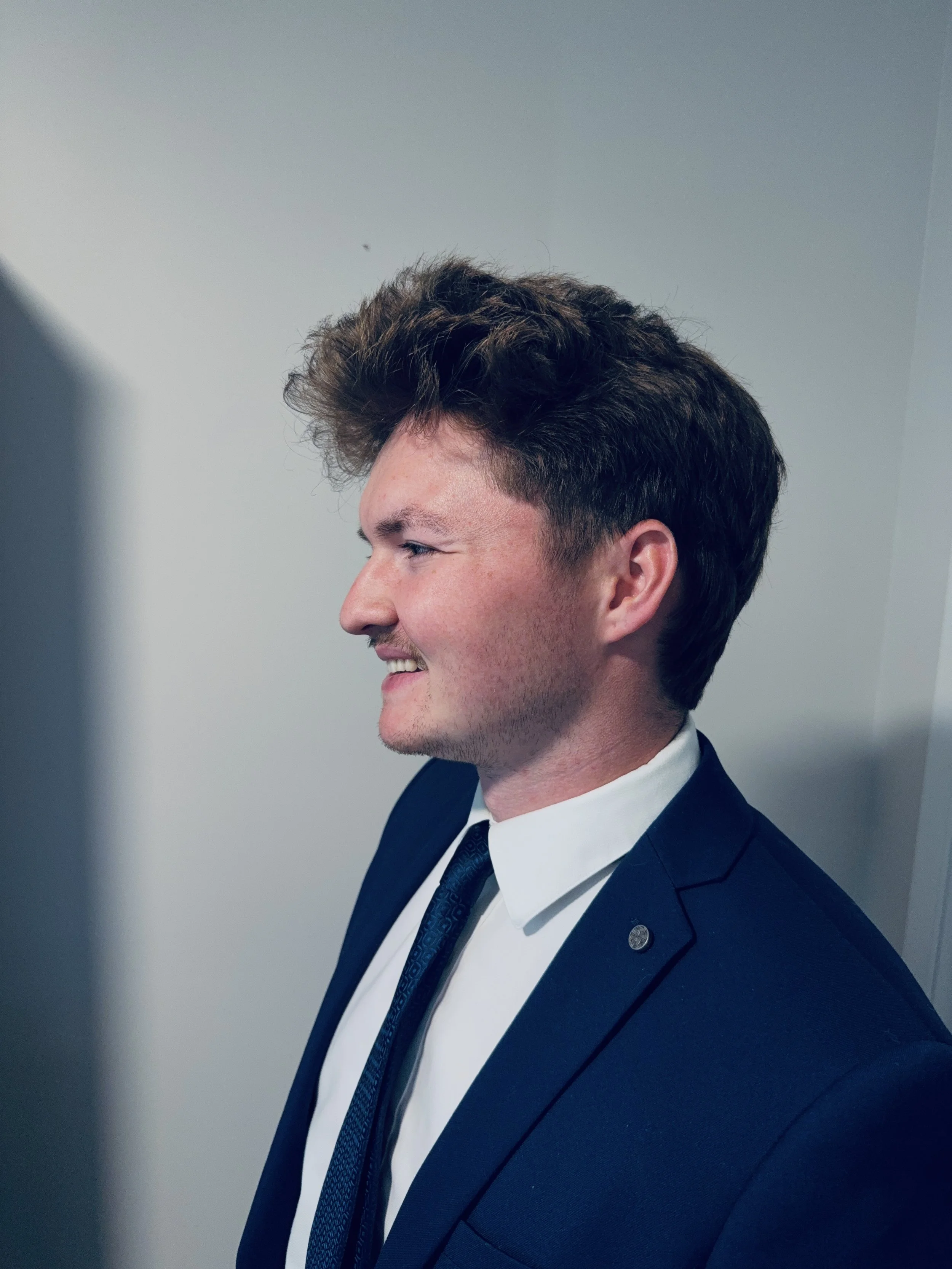 Side profile of a man wearing a dark blue suit, white shirt, and dark tie, smiling, with light-colored walls in the background.
