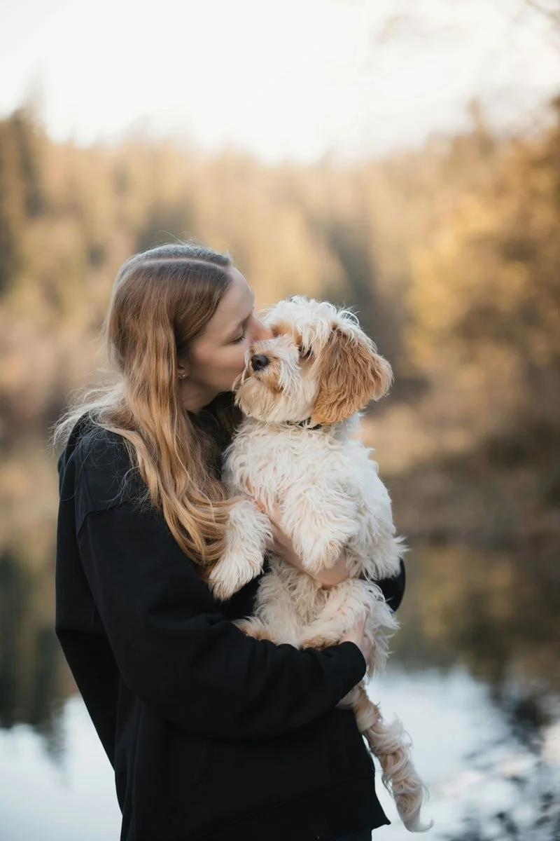 A woman holding and kissing a fluffy, light-colored dog outdoors with blurred trees and water in the background.