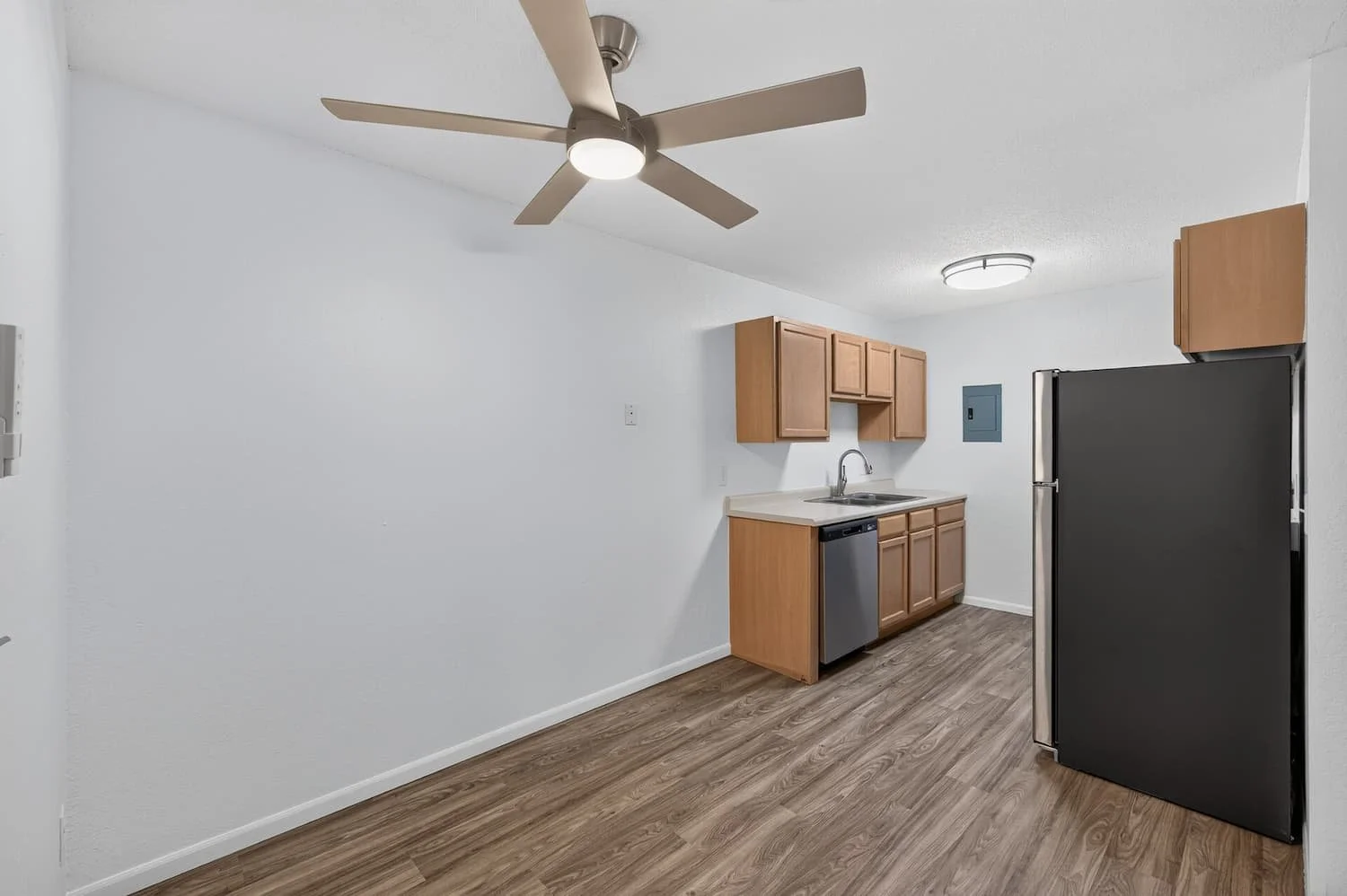 Empty kitchen with wooden cabinets, stainless steel dishwasher, black refrigerator, ceiling fan, and light fixture.