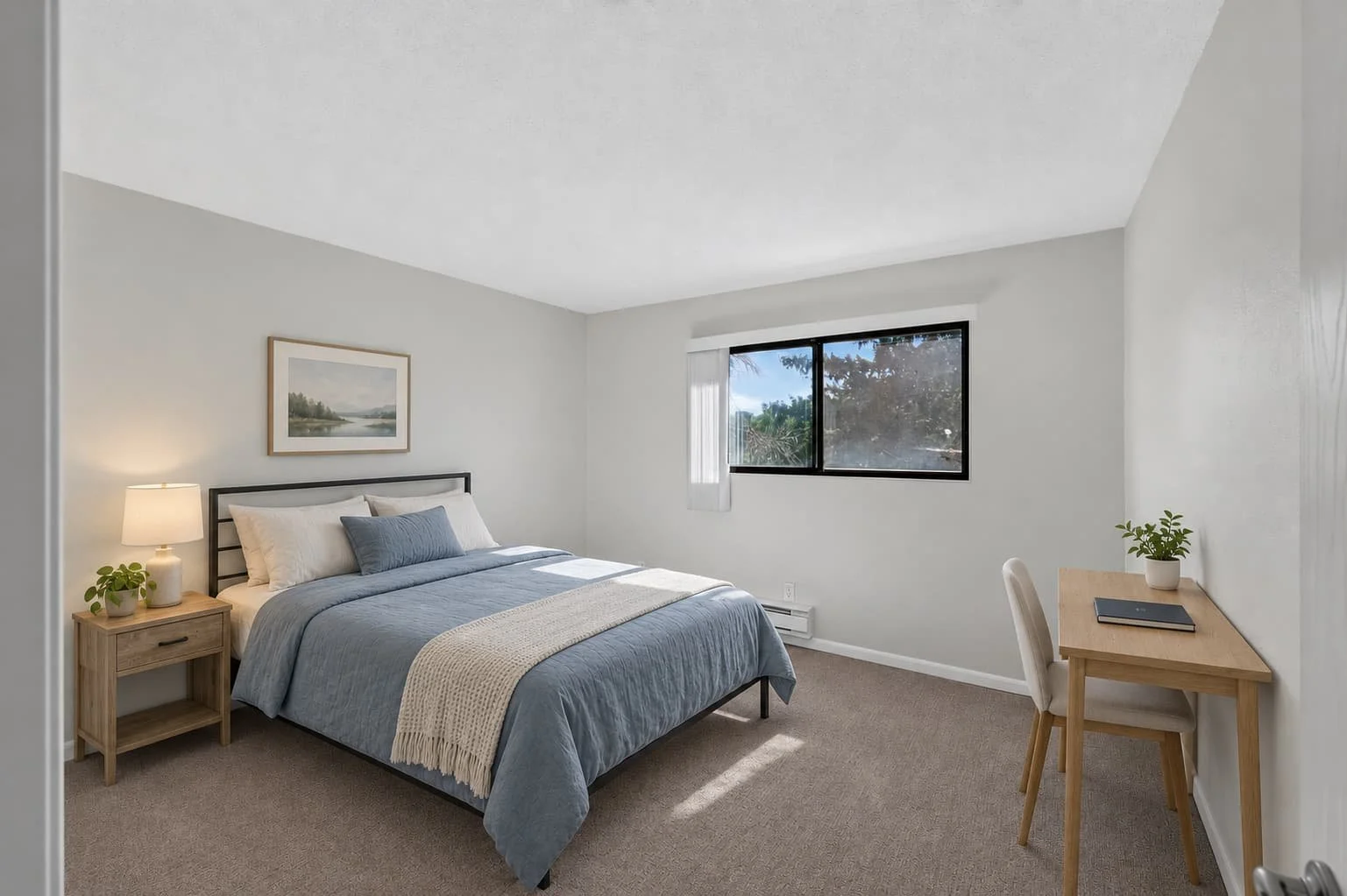 A bedroom with a bed covered in blue bedding, a beige blanket, and pillows, a bedside table with a lamp and plant, a window with white curtains, and a small wooden desk with a chair, plant, and laptop.