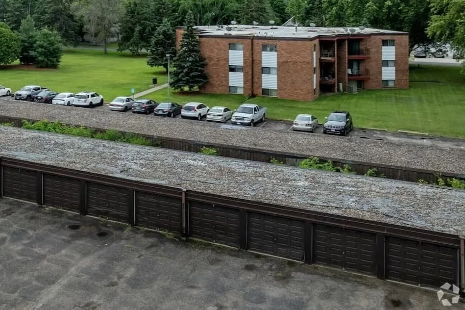 View of a row of parked cars behind a wooden fence, with an apartment building and green lawn and trees in the background.