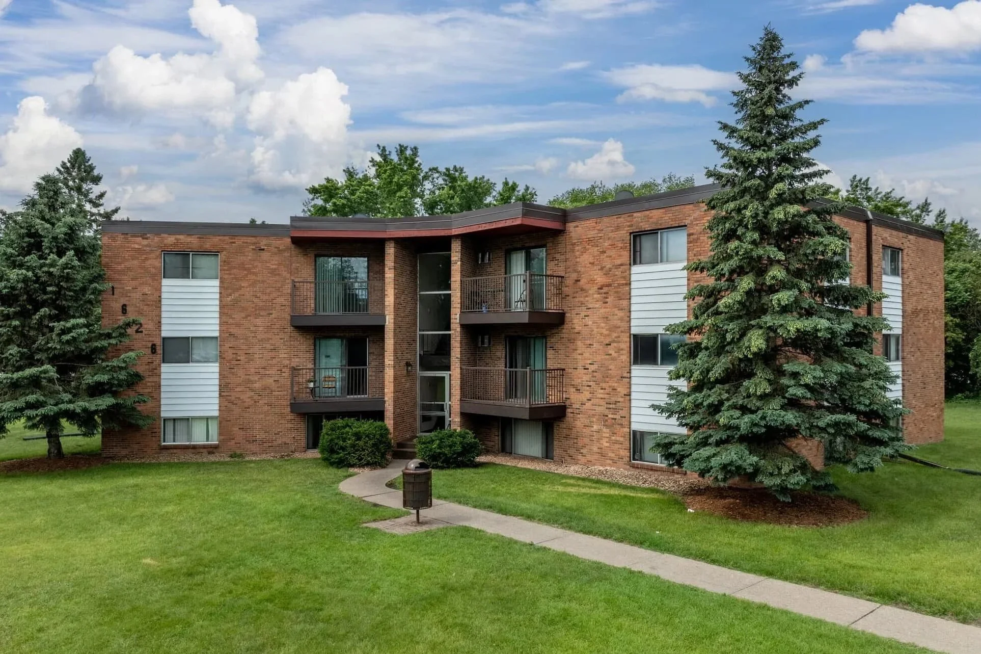 A three-story brick apartment building with multiple balconies, surrounded by green trees and grass, under a partly cloudy sky at Pineview Apartments in White Bear Lake
