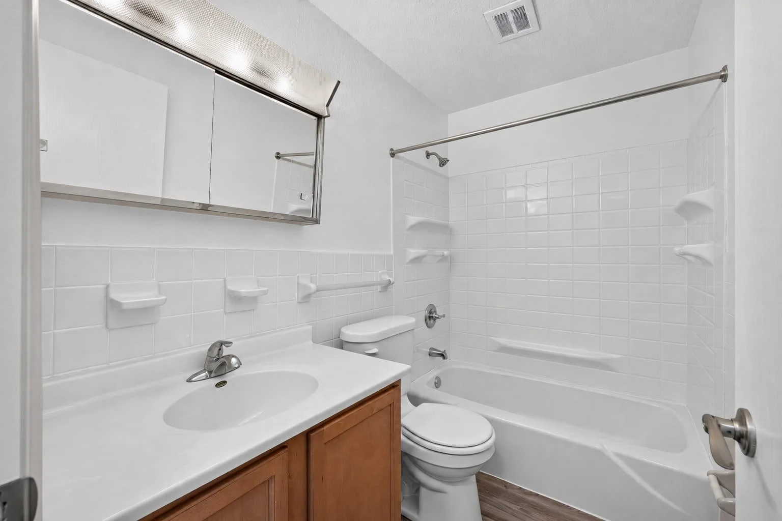 A bathroom with a white bathtub and shower, a toilet, a laminate wood vanity with a sink, a mirror, and white tiled walls.