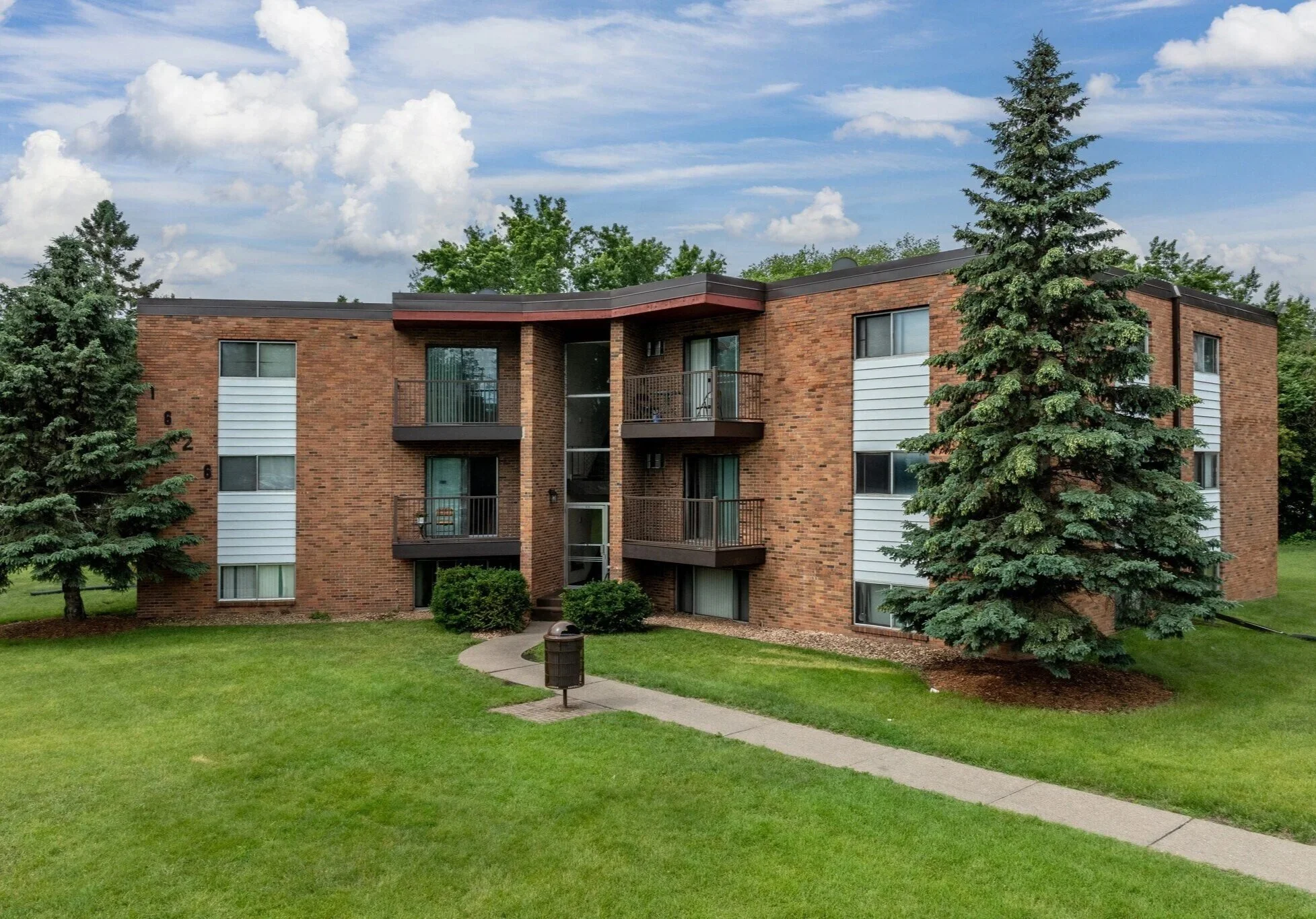 A multi-story brick apartment building with balconies, surrounded by tall evergreen trees and a well-maintained green lawn.