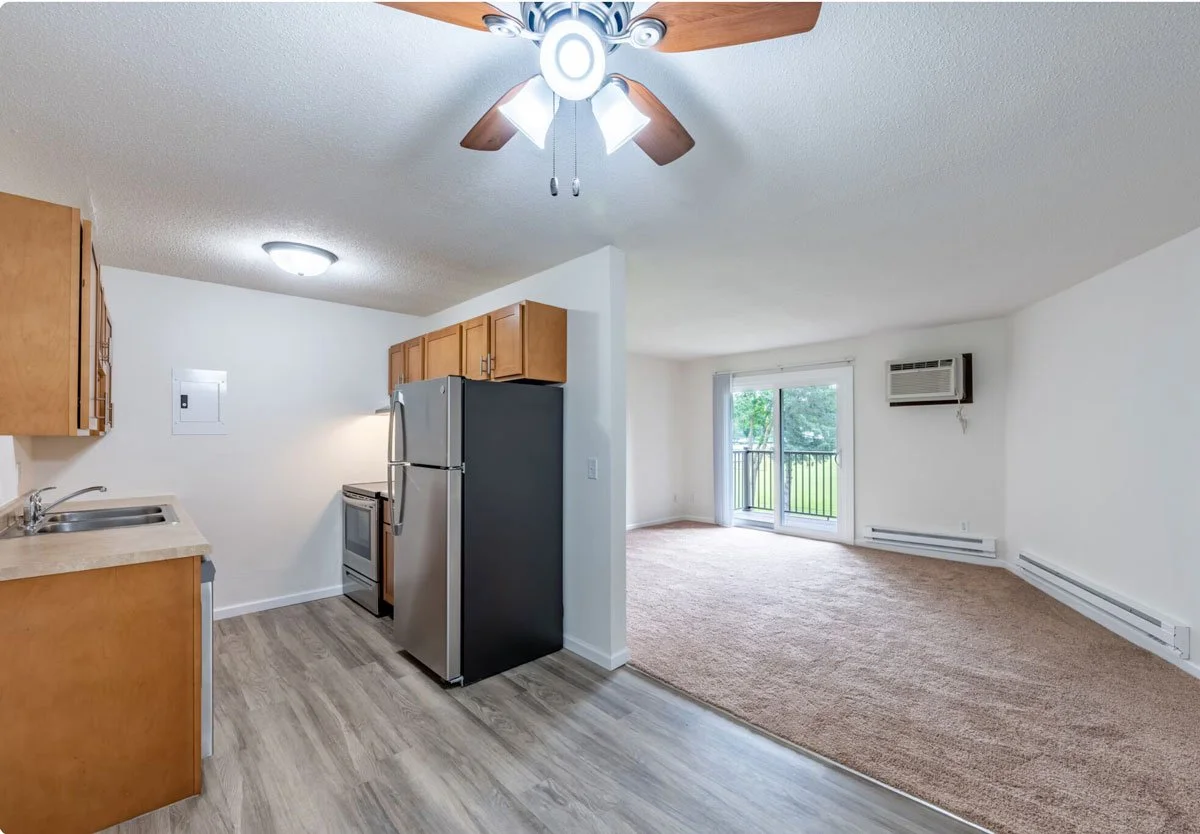 A living room and kitchen area in an apartment with sliding glass doors leading to a balcony, an air conditioning unit, and a ceiling fan with lights.