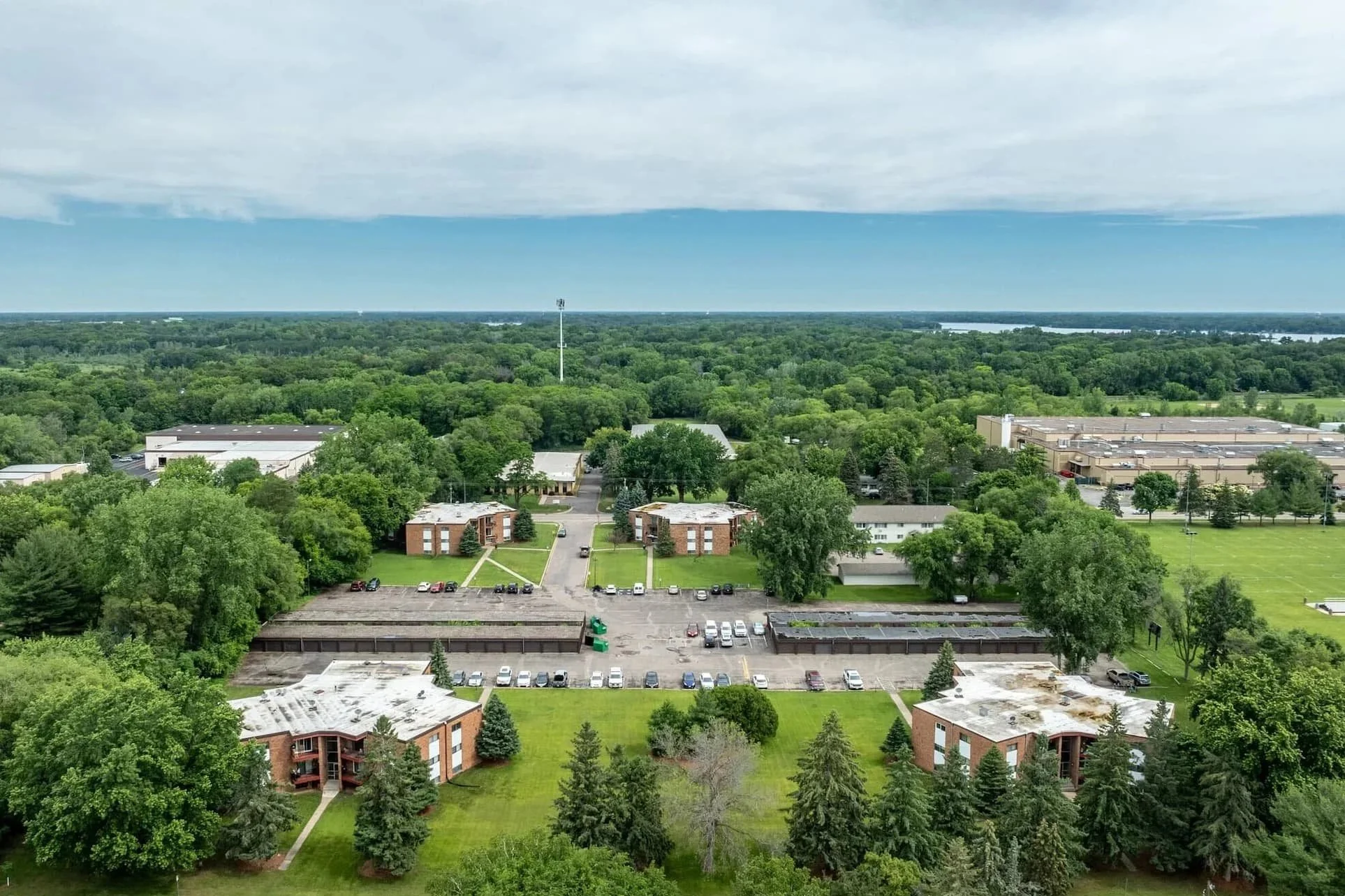 Aerial view of Pineview Apartments in White Bear Lakewith  a green, tree-filled landscape with a mix of residential and commercial buildings, parking lots, and a water body in the distance under a cloudy sky.