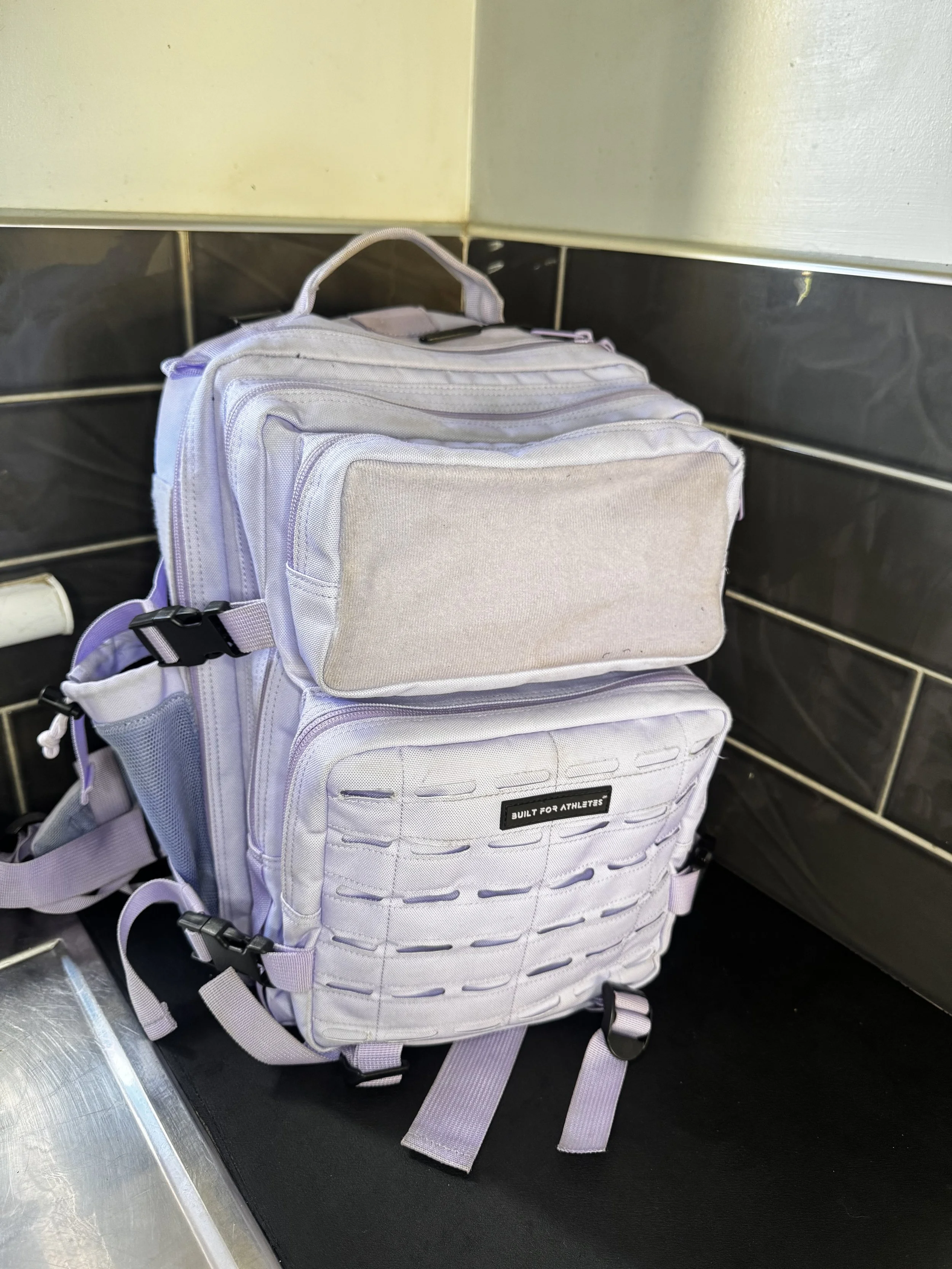 Light purple backpack with multiple compartments placed on a black surface against a tiled wall.