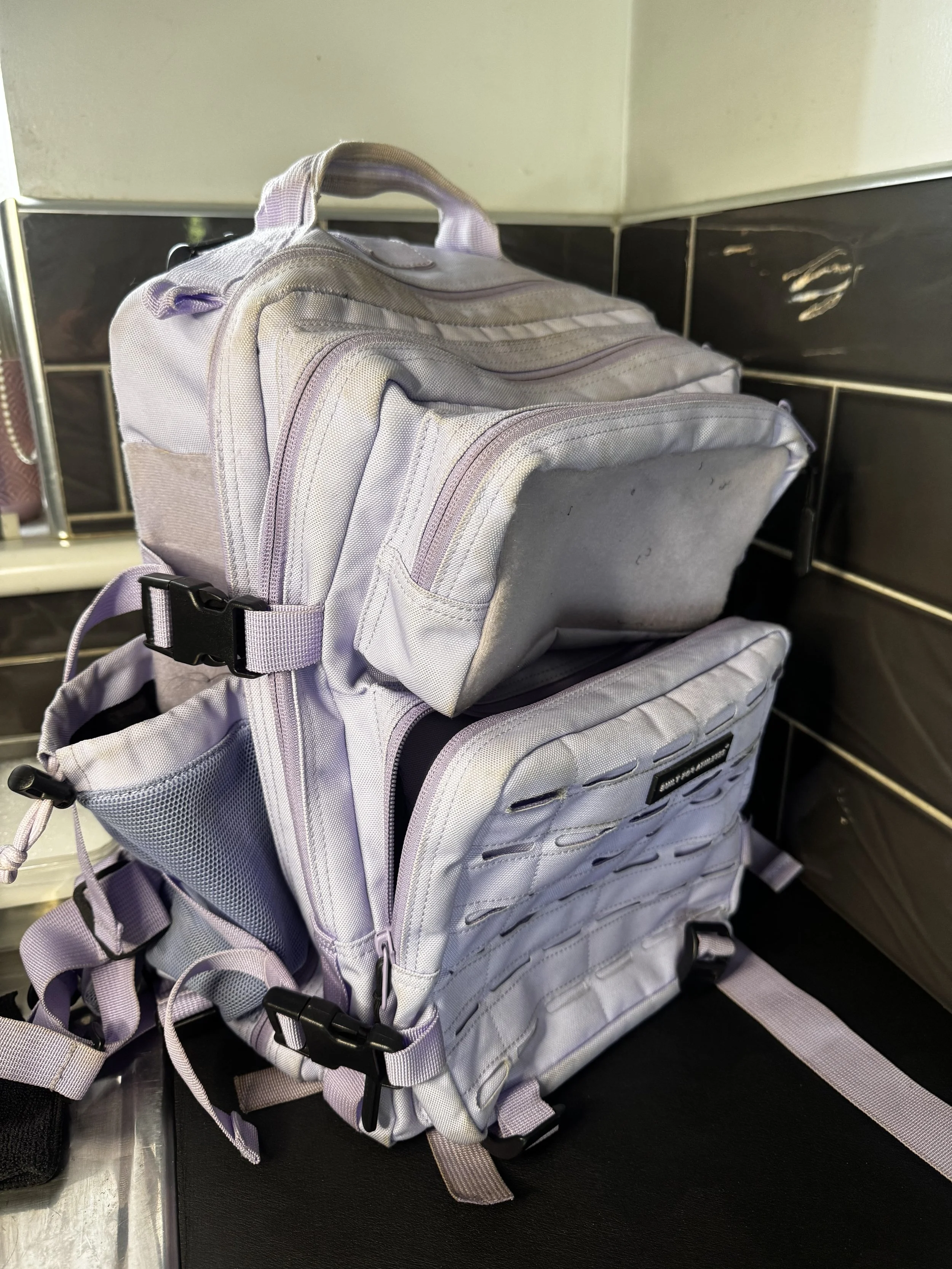 A light gray backpack with multiple compartments and adjustable straps, placed on a black surface against a wall with black and beige tiles.