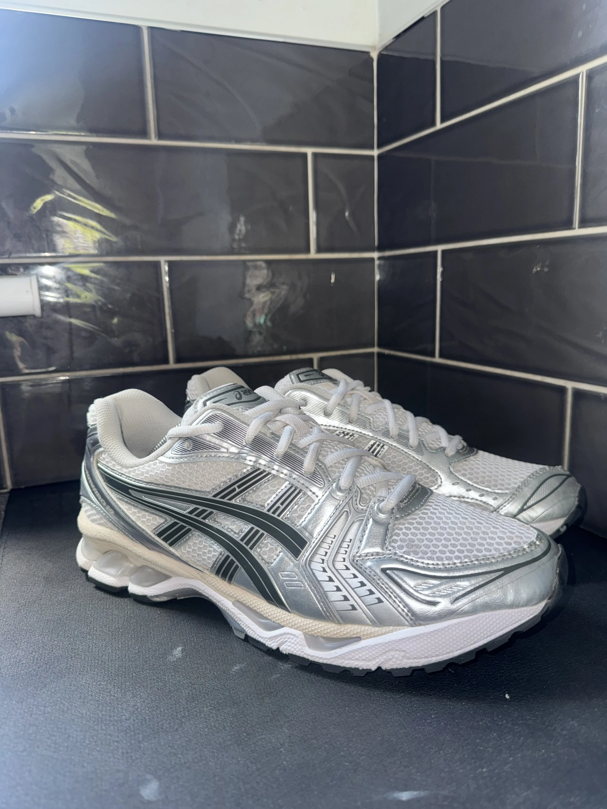 A pair of white and silver athletic shoes with black accents placed on a black surface in front of black tiled walls.