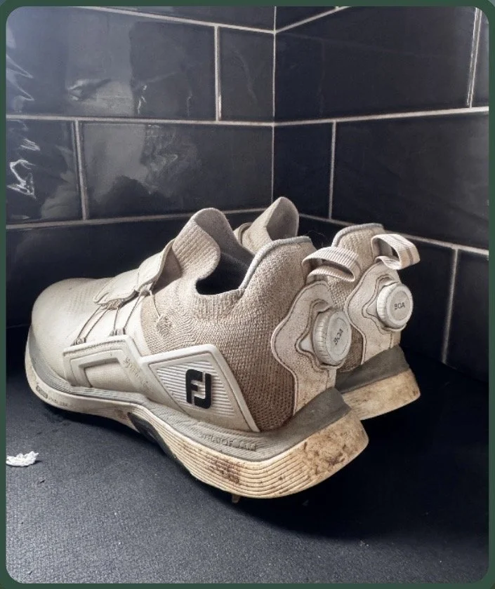 A pair of white FootJoy golf shoes with BOA dial laces, sitting on a dark surface against dark tiled wall, with visible dirt and wear on the soles.