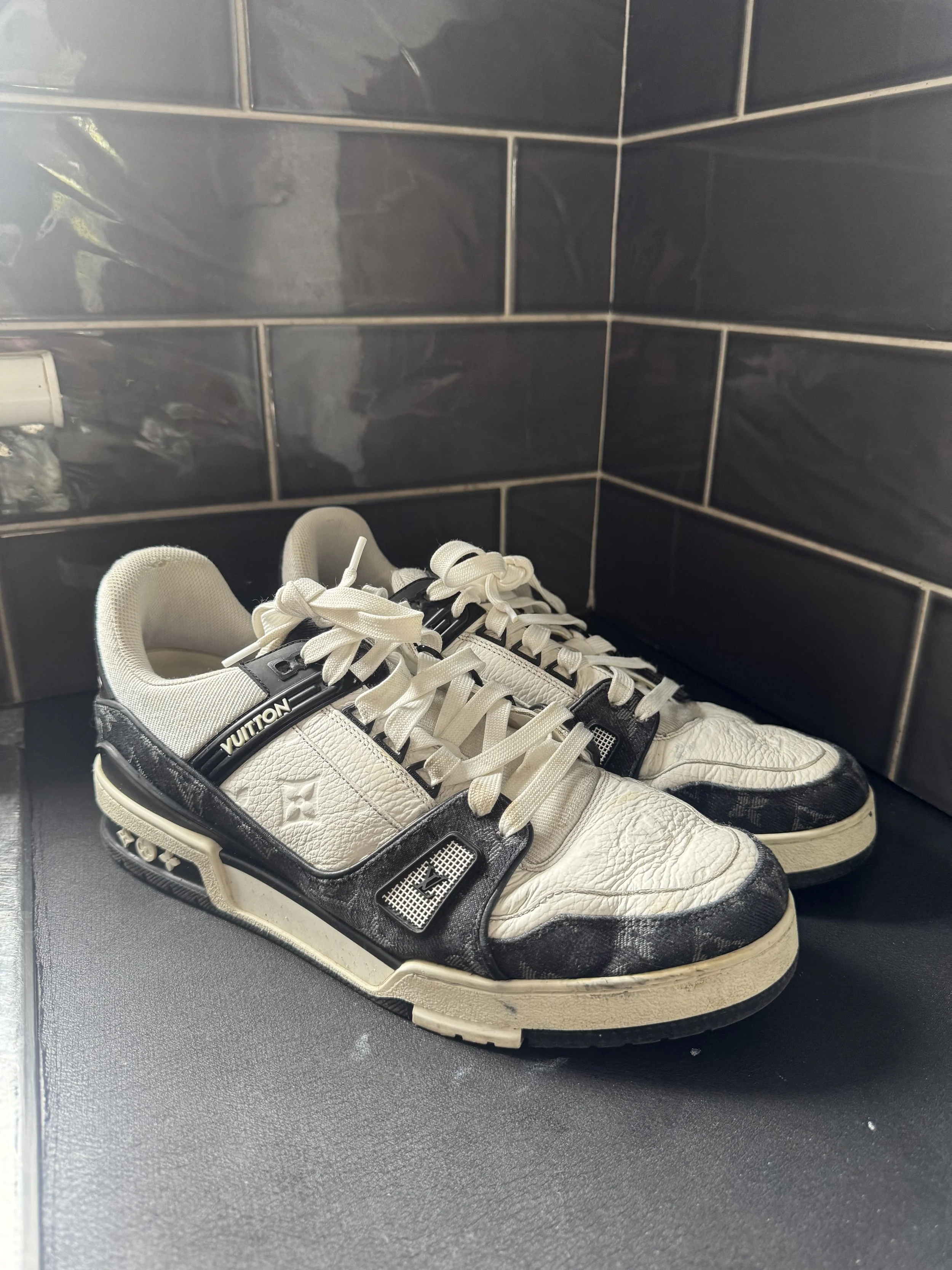 A pair of white and black Louis Vuitton sneakers with a chunky sole positioned on a black surface against a black tiled wall background.