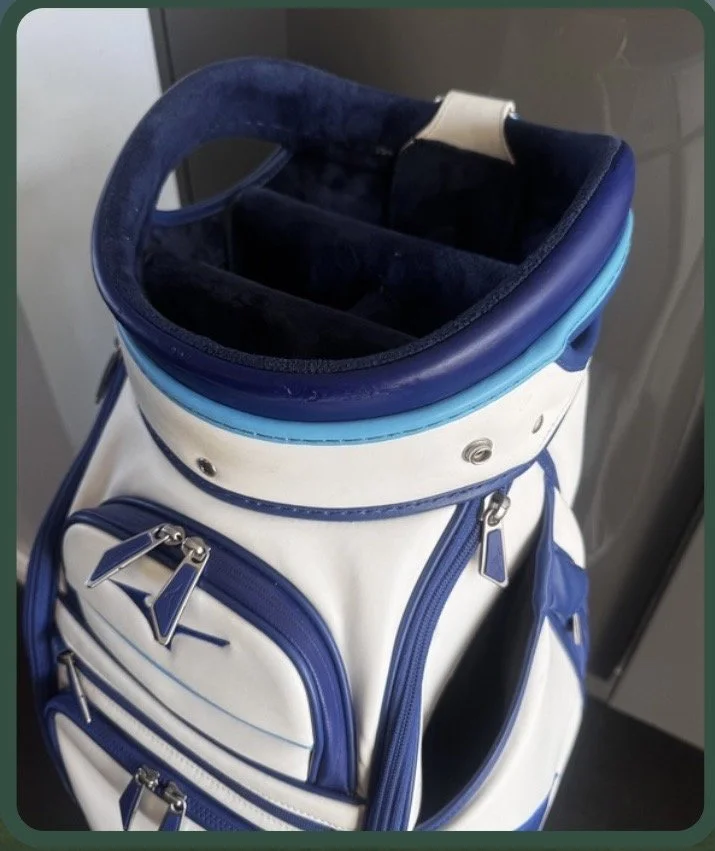 A white and blue golf bag with multiple zippered compartments standing on a floor.