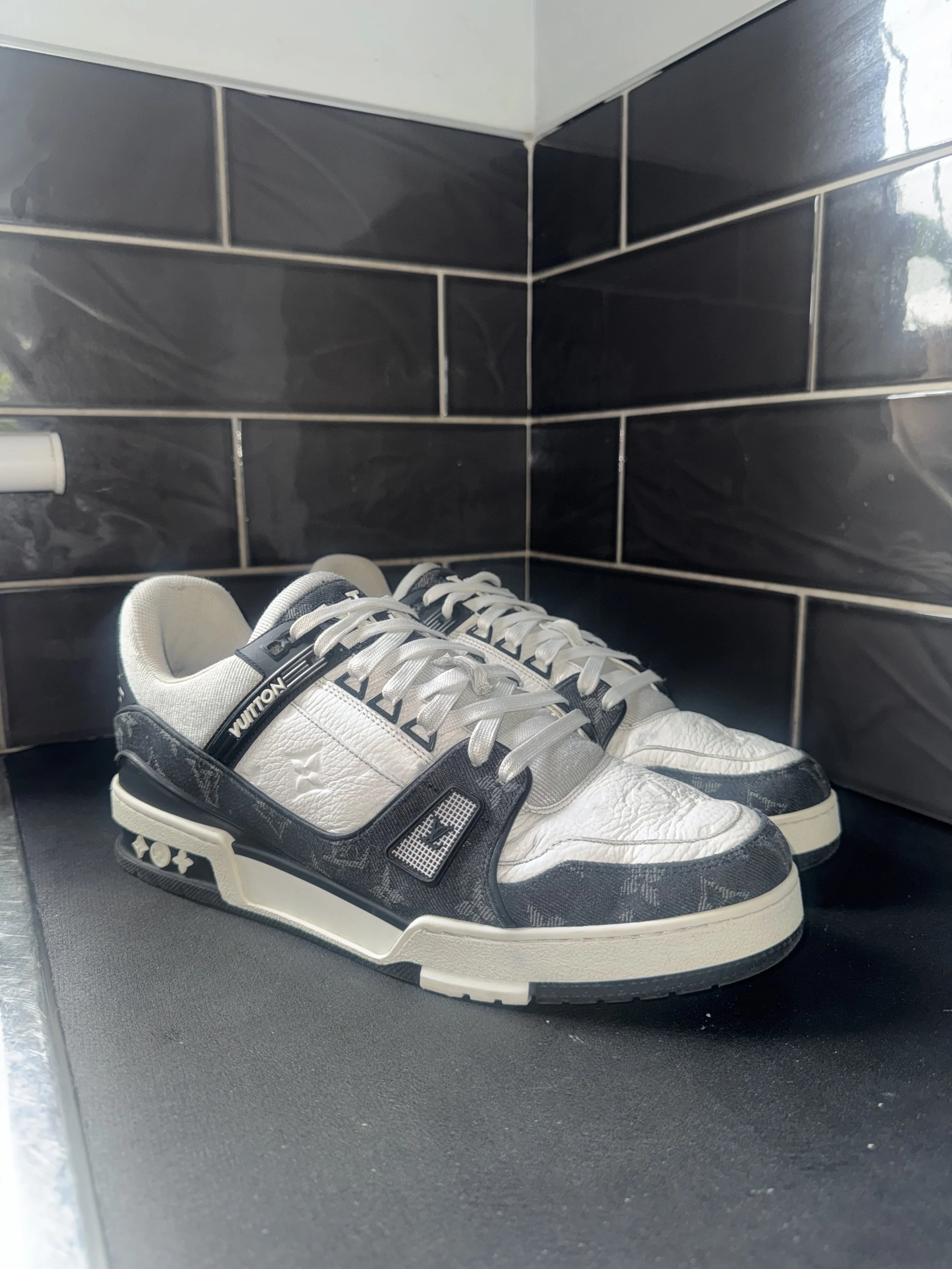 A pair of white and black sneakers resting on a dark tiled floor, with part of a white wall and a plumbing pipe visible in the background.