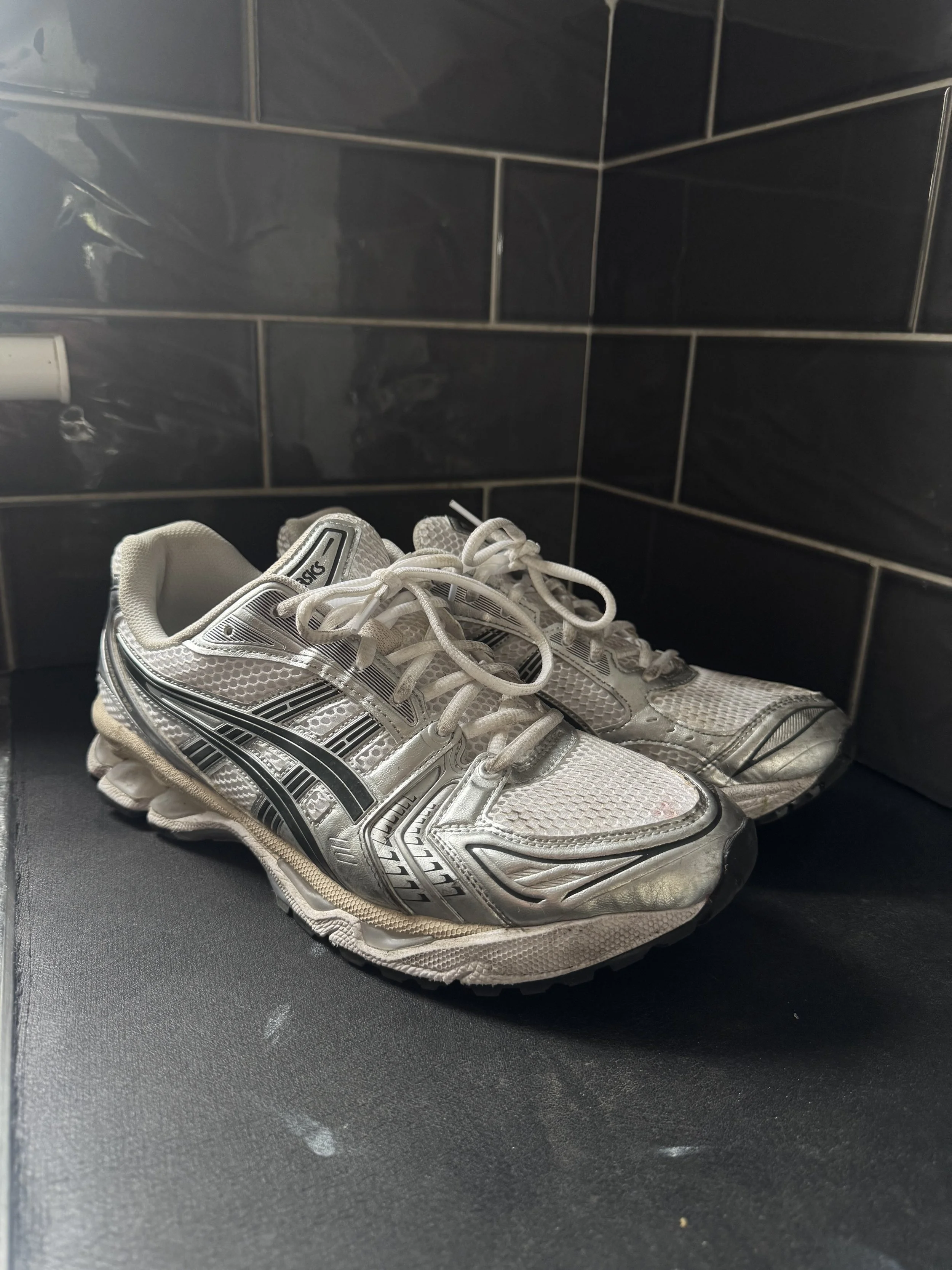 A pair of white and silver athletic shoes on a black surface with black tiled walls in the background.
