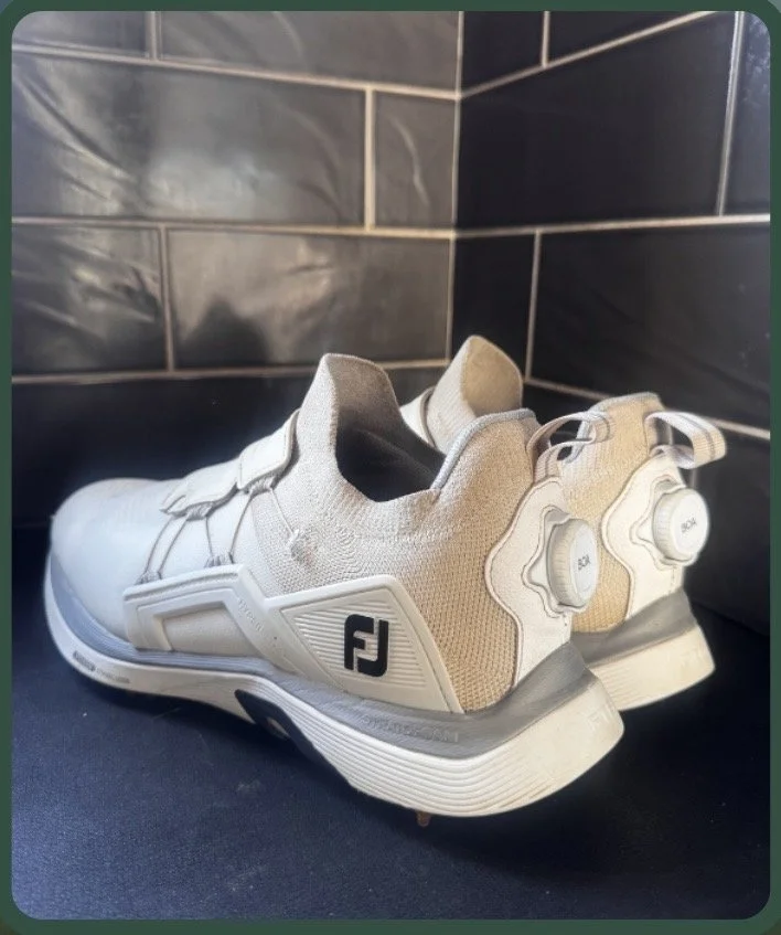 White and beige FootJoy golf shoes on a black surface in front of black tiled wall.