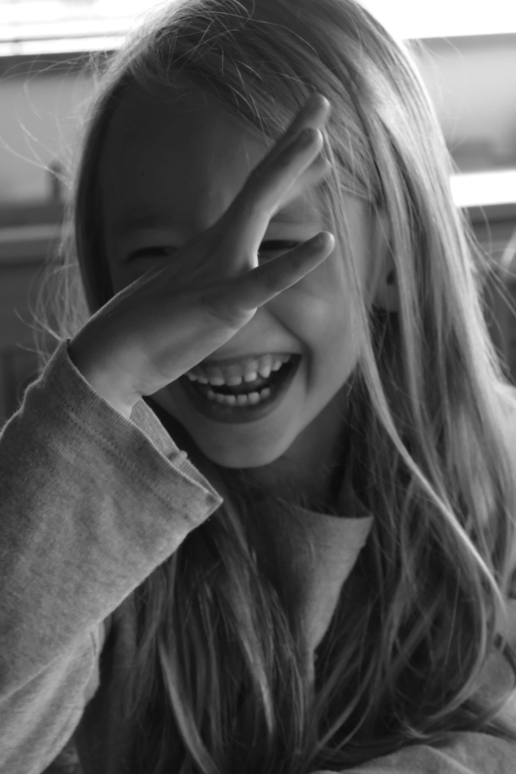 A girl with long hair covering part of her face in a playful pose, smiling and holding her hand near her eye.