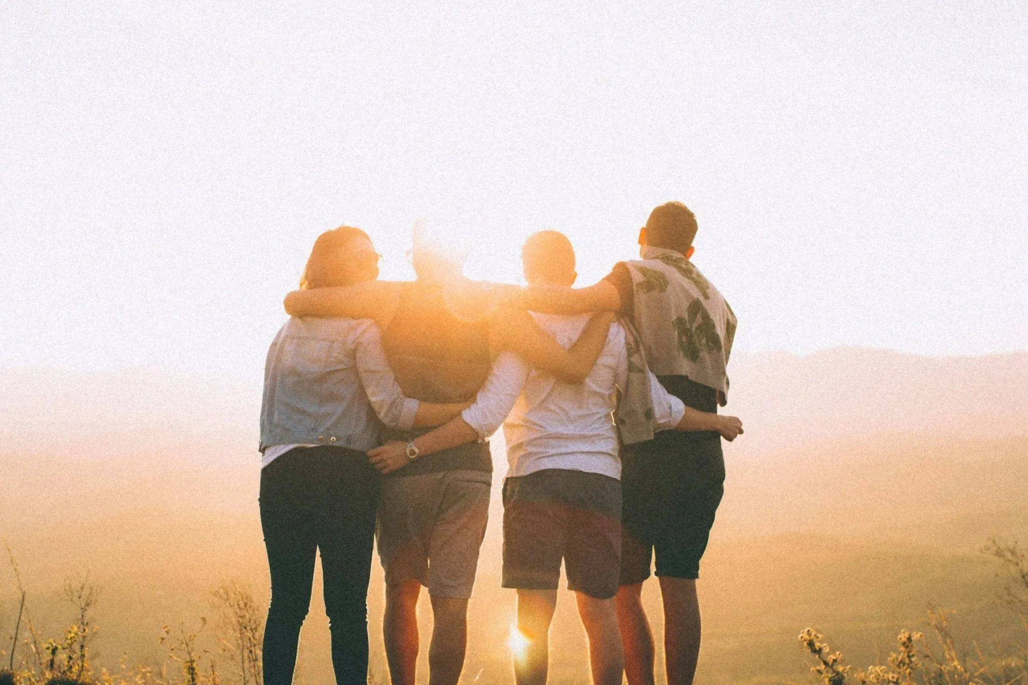Four friends standing close together during sunset, arms around each other, enjoying the moment.