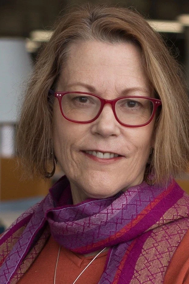 A middle-aged woman with shoulder-length wavy hair wearing red glasses, a purple patterned scarf, and an orange top.