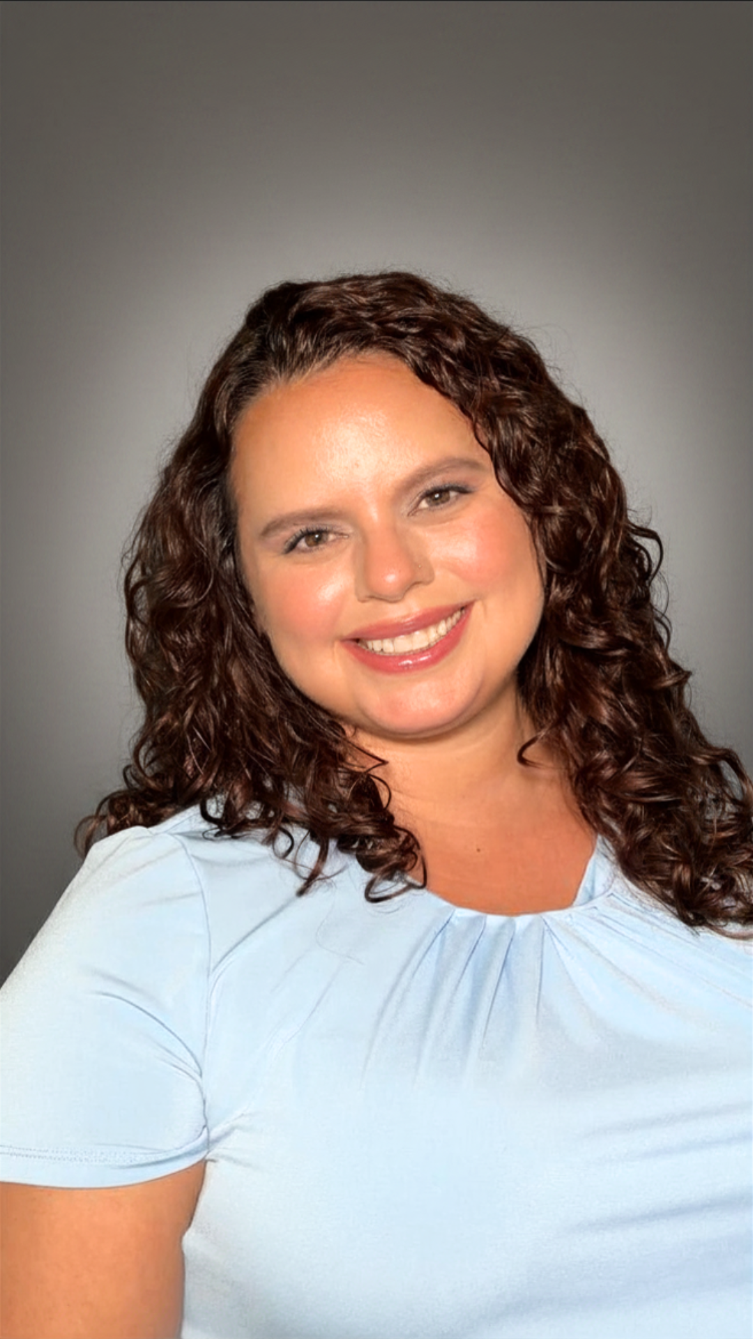 A smiling woman with curly brown hair wearing a light blue top against a gray background.