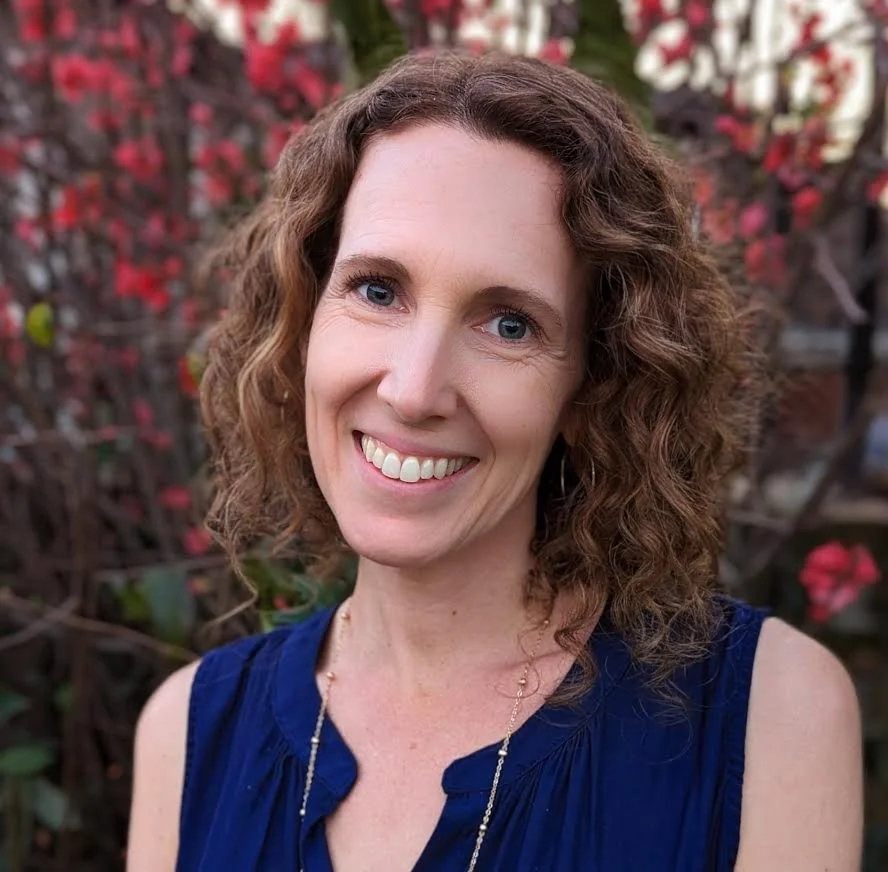 A woman with shoulder-length curly brown hair and blue eyes smiling outdoors in front of a red and green foliage background, wearing a blue sleeveless top and a delicate necklace.