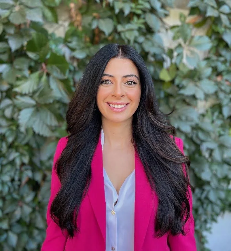 A woman with long dark hair, wearing a pink blazer over a white blouse, standing in front of green leafy foliage, smiling at the camera.
