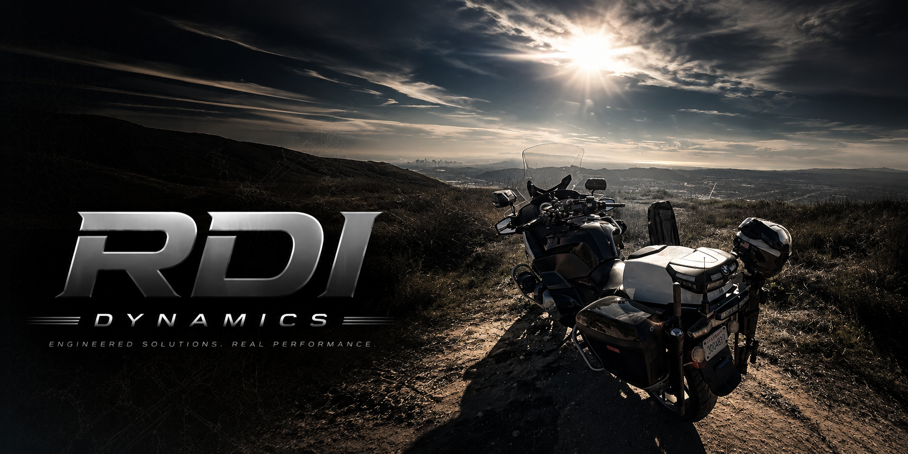 A black motorcycle with a windshield and a helmet attached, parked on a dirt trail in a hilly landscape during sunset. The sky is partly cloudy with the sun low on the horizon. The image includes the text 'RDI DYNAMICS' and 'Engineered solutions. Real performance.' in the lower left corner.