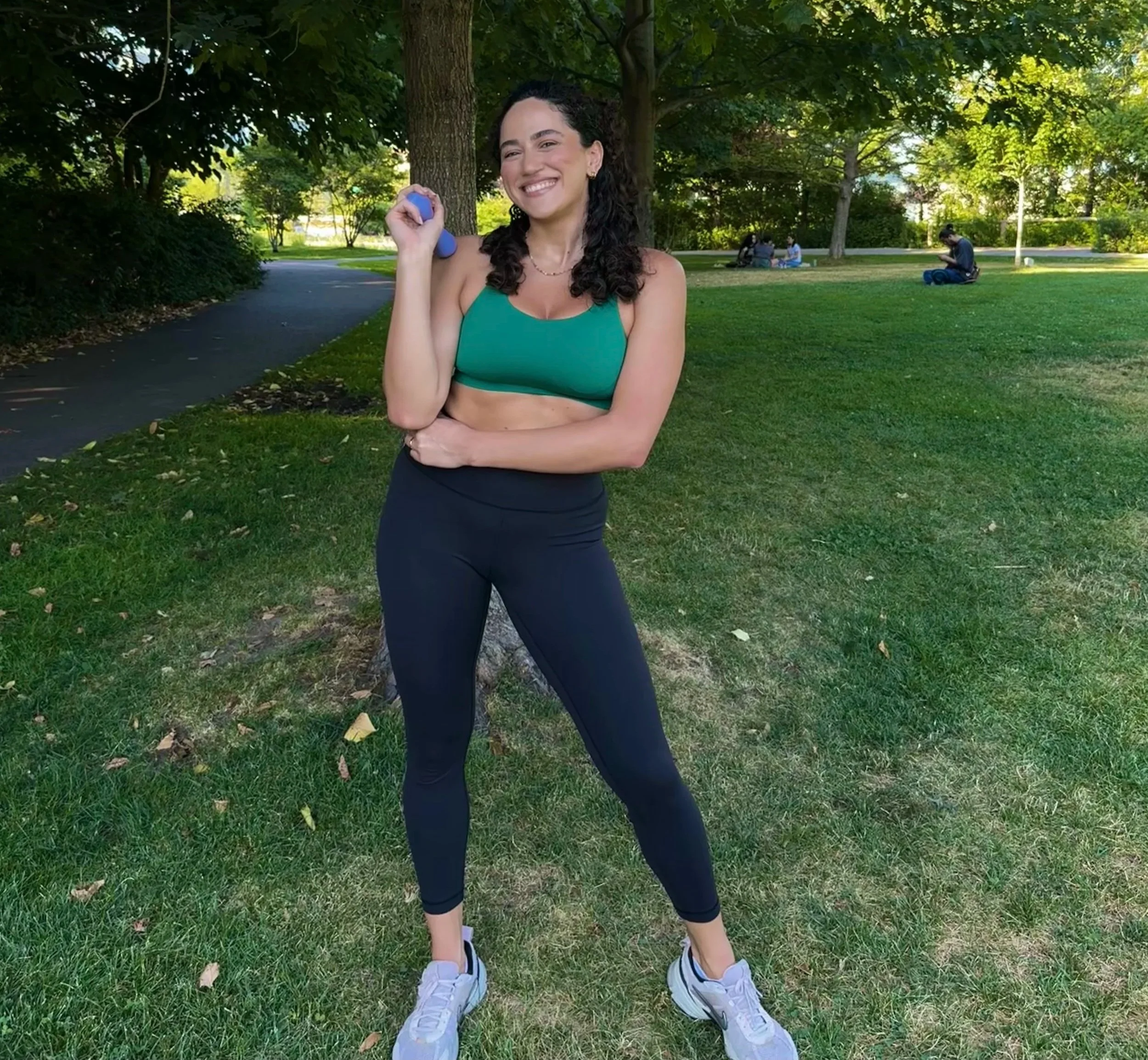 Young woman holding small dumbbell outdoors in a park with trees and grass, smiling happily.