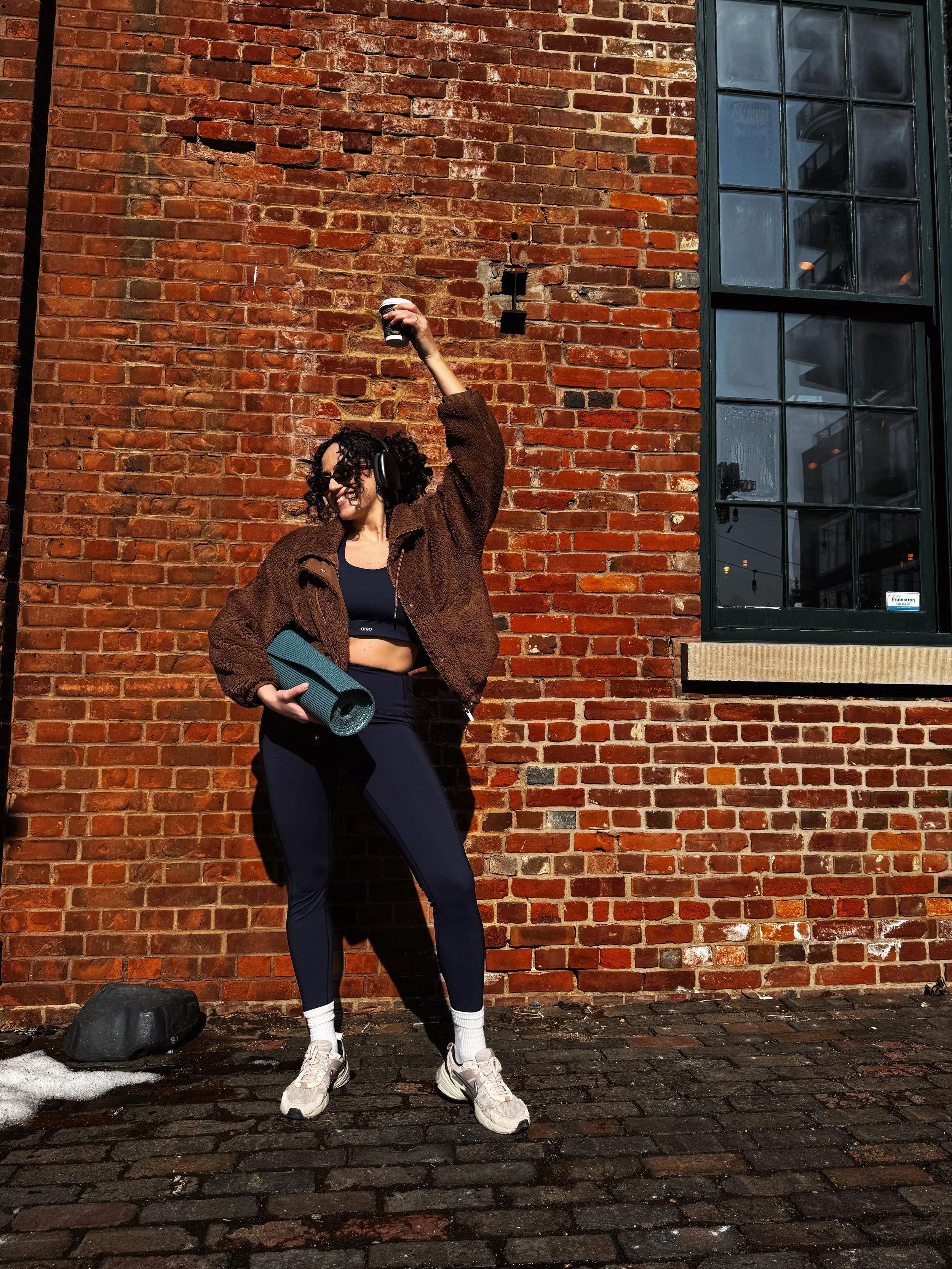 Woman with curly hair, sunglasses, and headphones, holding a yoga mat and a coffee cup, standing against a brick wall on a cobblestone street.