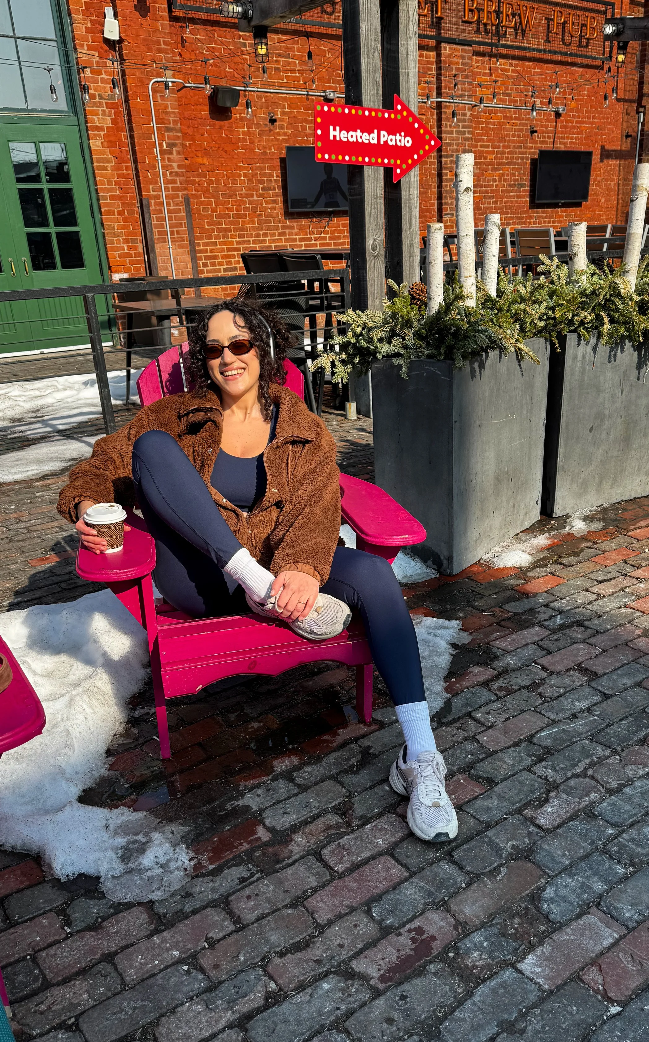 A woman sitting on a pink chair outdoors next to a snow patch, holding a coffee cup, wearing sunglasses, a brown jacket, navy leggings, and white sneakers with socks, smiling in front of a brick building with a sign pointing to a heated patio.