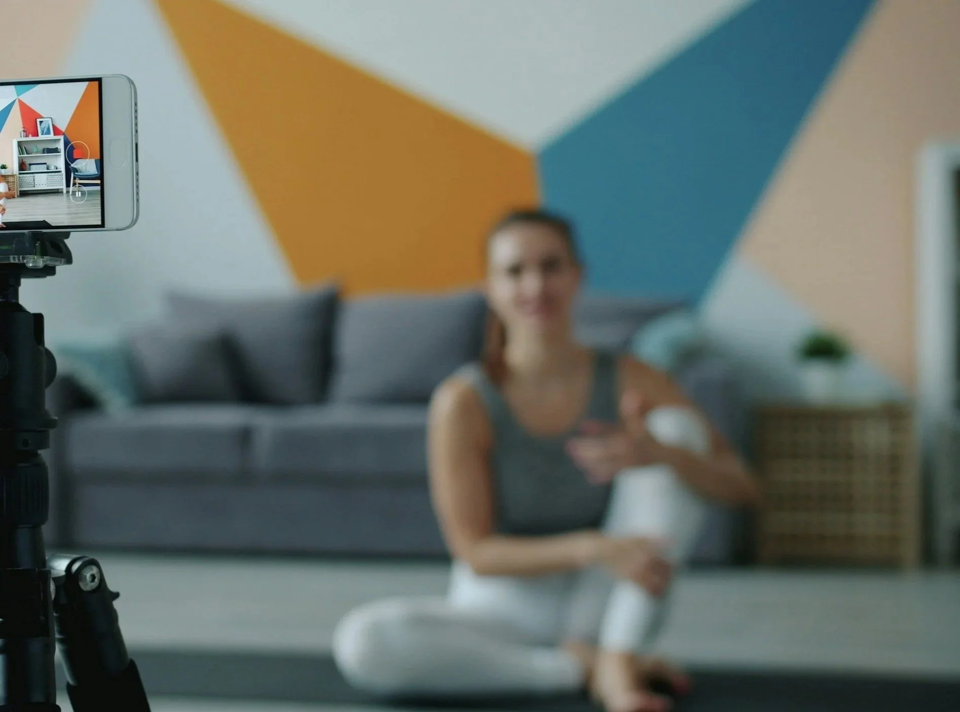 A blurred person sitting on a couch in front of a colorful geometric wall, with a smartphone mounted on a tripod capturing the scene.