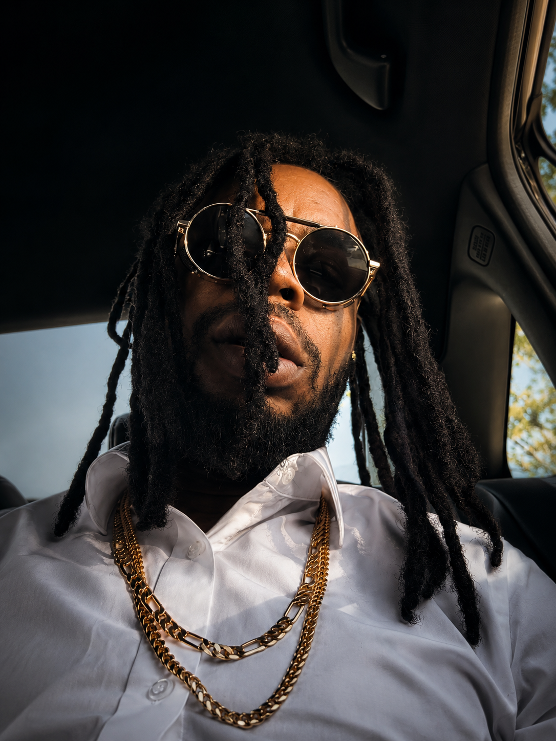 Close-up of a man with dreadlocks and sunglasses, wearing a white shirt and gold chains, sitting inside a car with sunlight and trees visible outside.