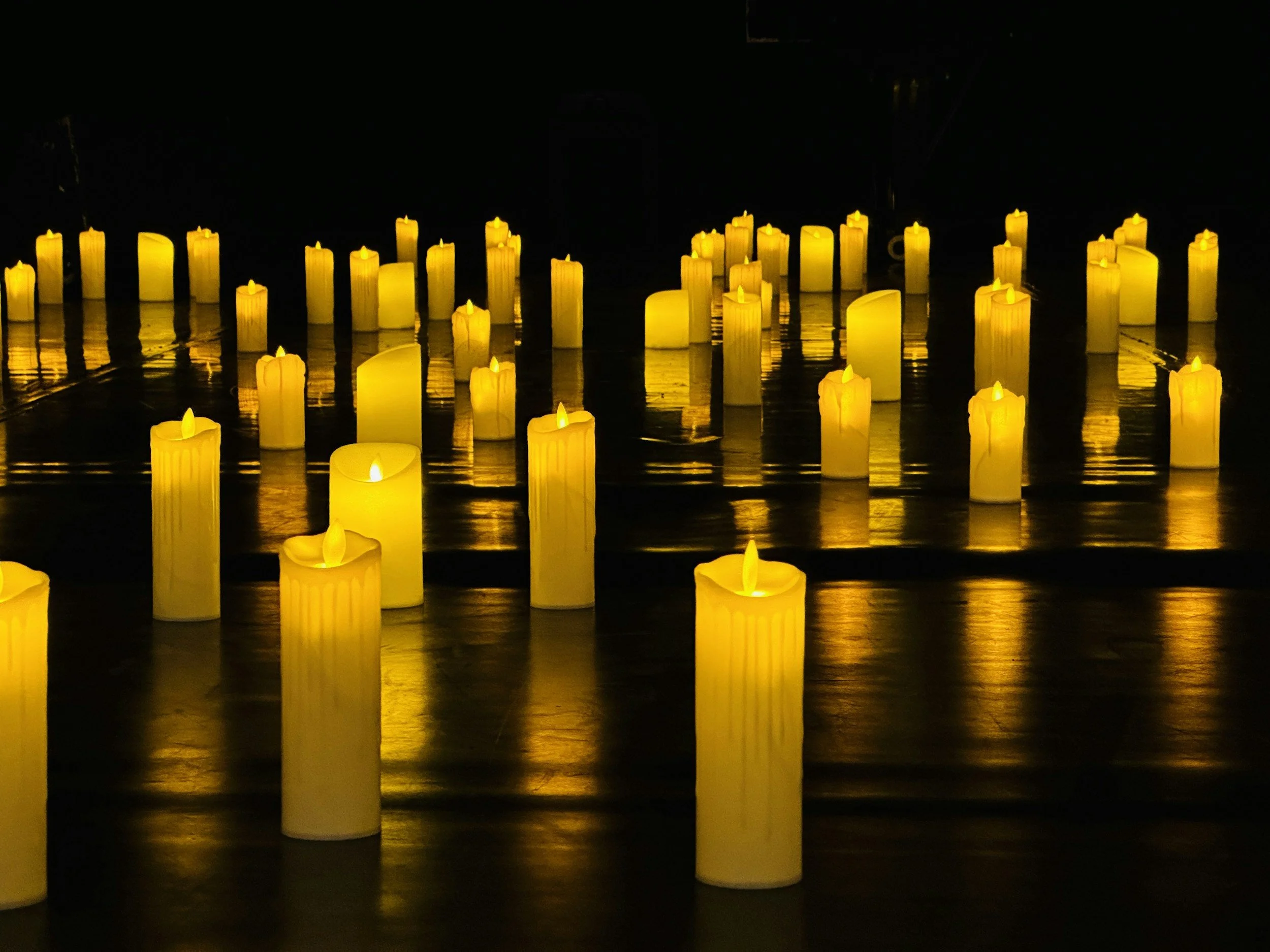 Numerous yellow candles placed on a reflective black surface, some partially melted, glowing softly in the dark.