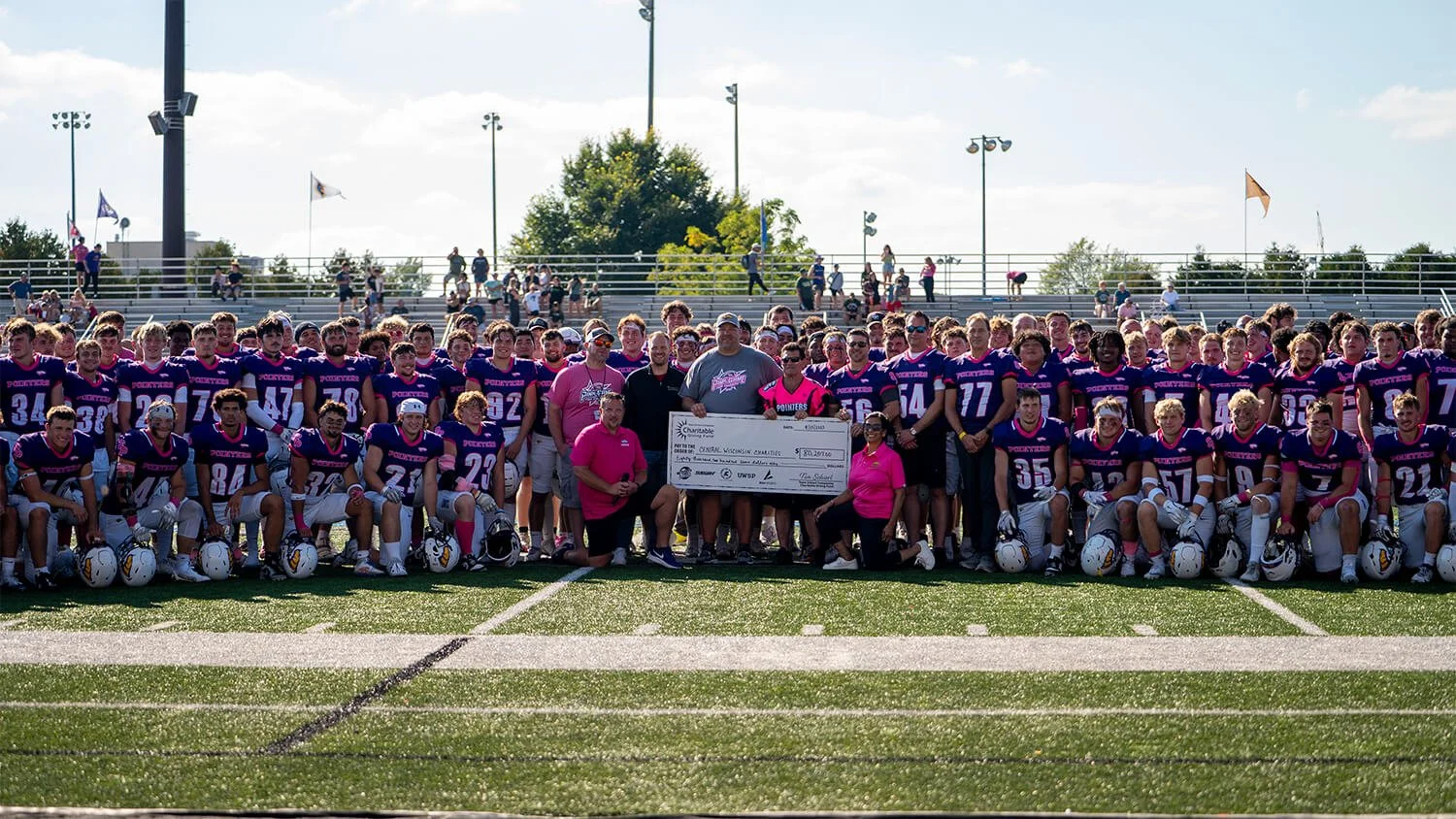 Home and rival teams posing together at the Pink Game for Charity, along with a massive check showing money raised.