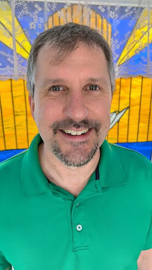 A smiling man with short brown hair, gray beard, wearing a green polo shirt, standing in front of a colorful, stained glass window with yellow, orange, blue, and purple sections.
