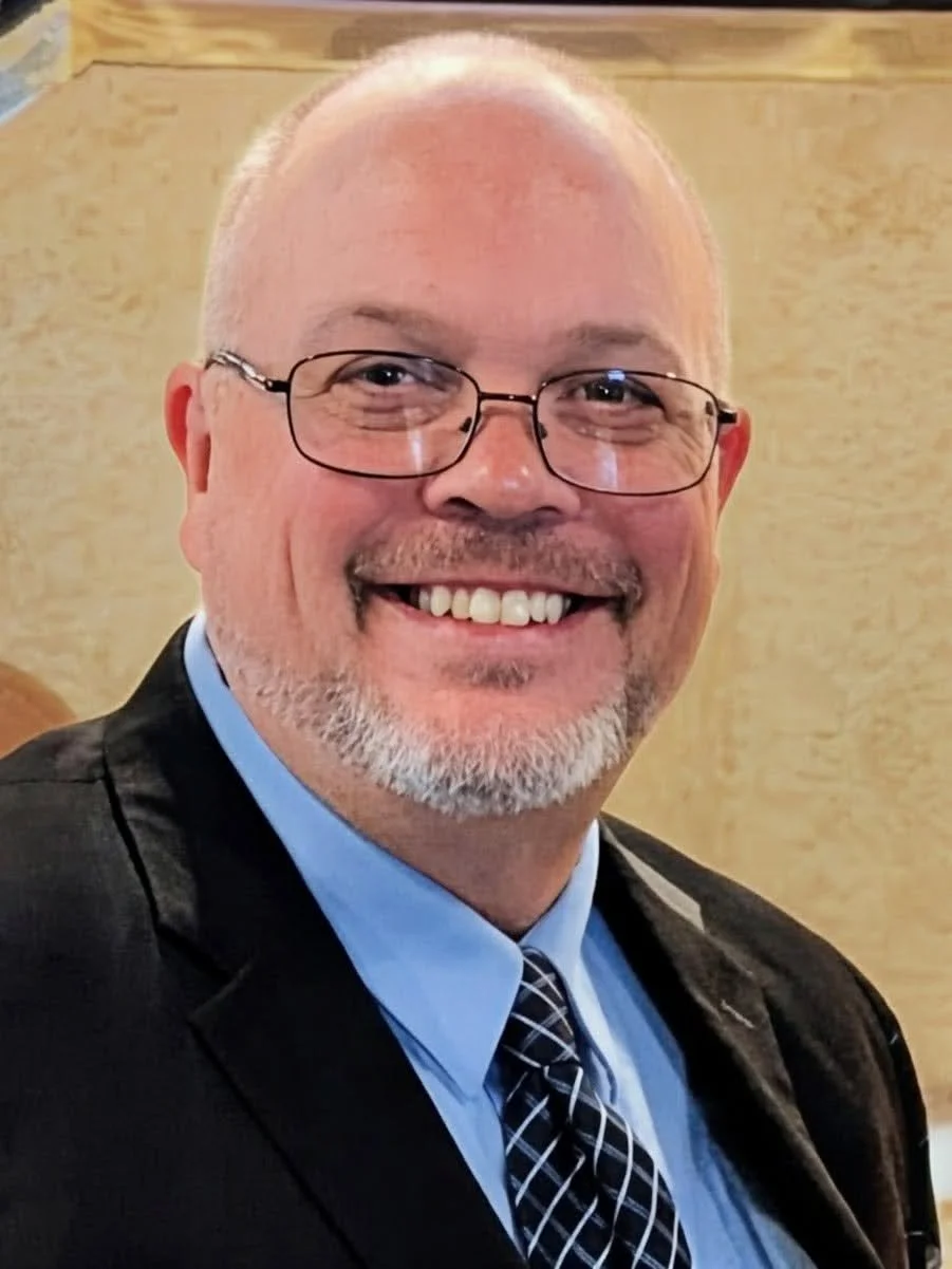 Close-up of smiling man wearing glasses, a suit, and a patterned tie, with a wooden background.