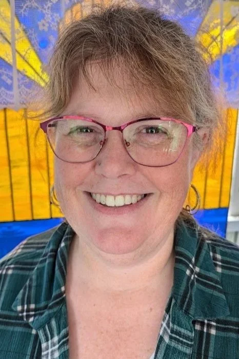A woman smiling outdoors, wearing pink glasses and a plaid shirt, with yellow and blue decorations in the background.