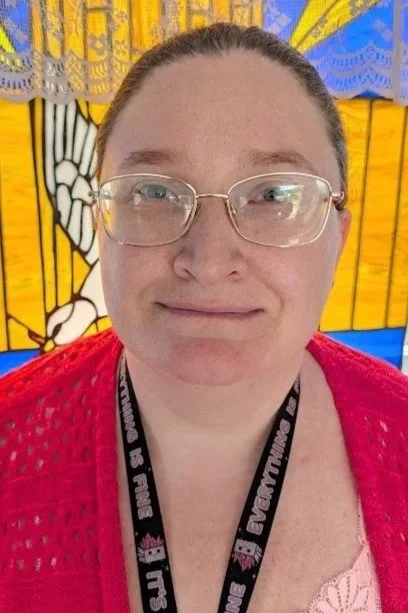 A woman with glasses and a red top standing in front of a colorful stained glass window.