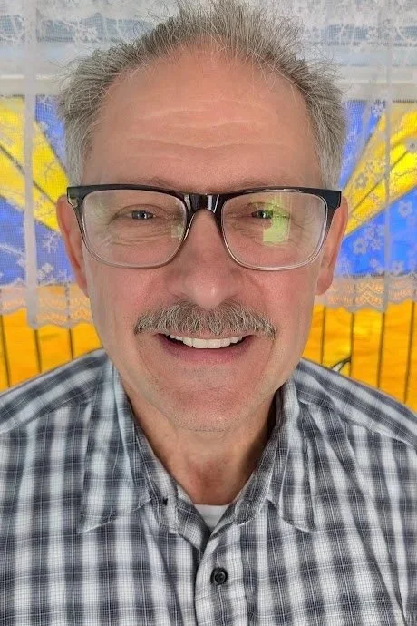 A smiling older man with glasses and a checkered shirt, standing indoors with yellow and blue decorations in the background.