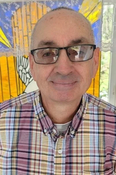 A smiling man with glasses and a checkered shirt standing indoors in front of a colorful stained glass window background.