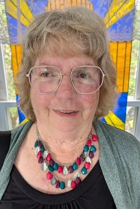 A smiling elderly woman with glasses and short gray hair, wearing a multicolored beaded necklace, in front of a stained glass window with blue and yellow pieces.