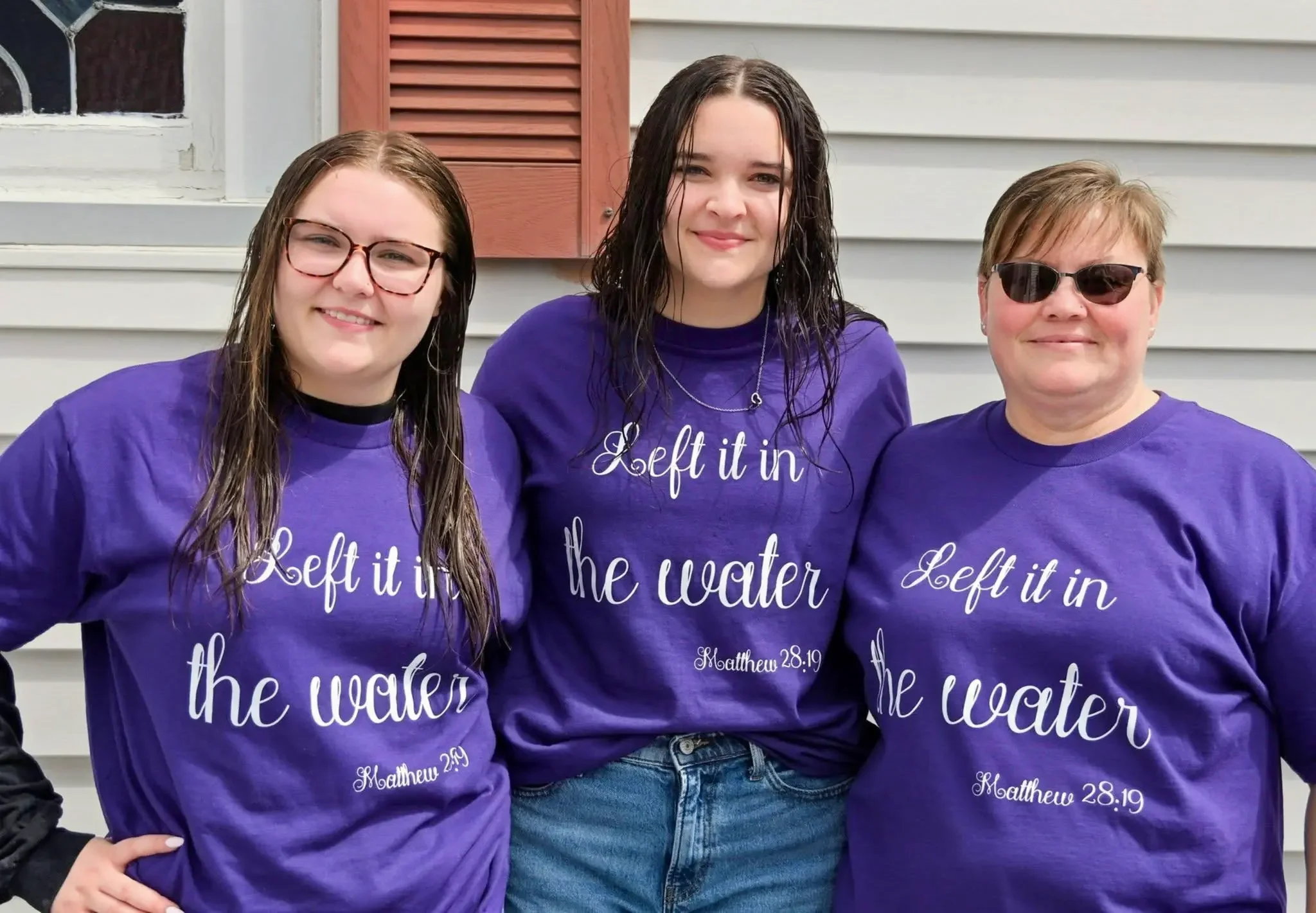Group of three women wearing matching purple shirts with white text that reads 'Left it in the water,' standing outdoors in front of a house with white siding and a window with brown shutters. The women are smiling.